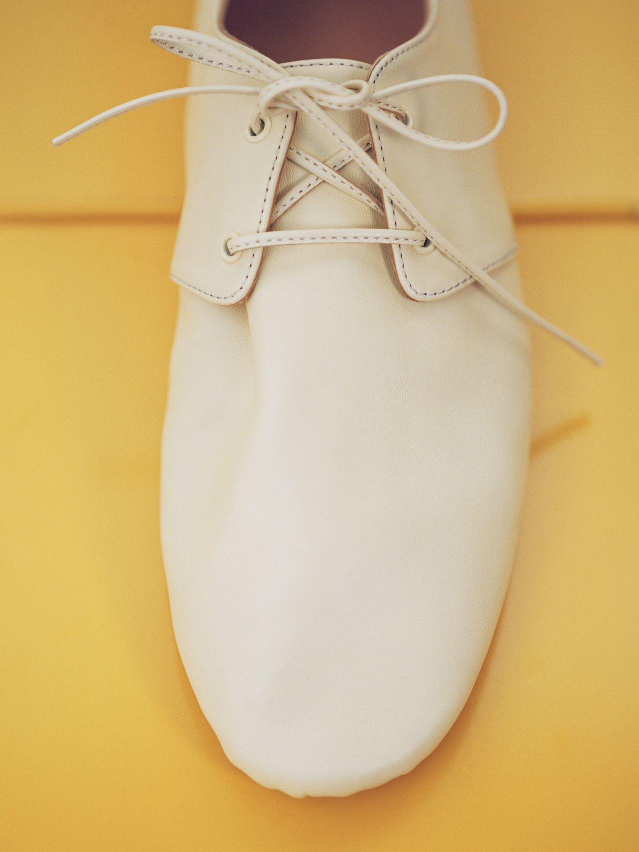 A cream-colored, soft-textured shoe on a yellow background, featuring white laces and a smooth design.