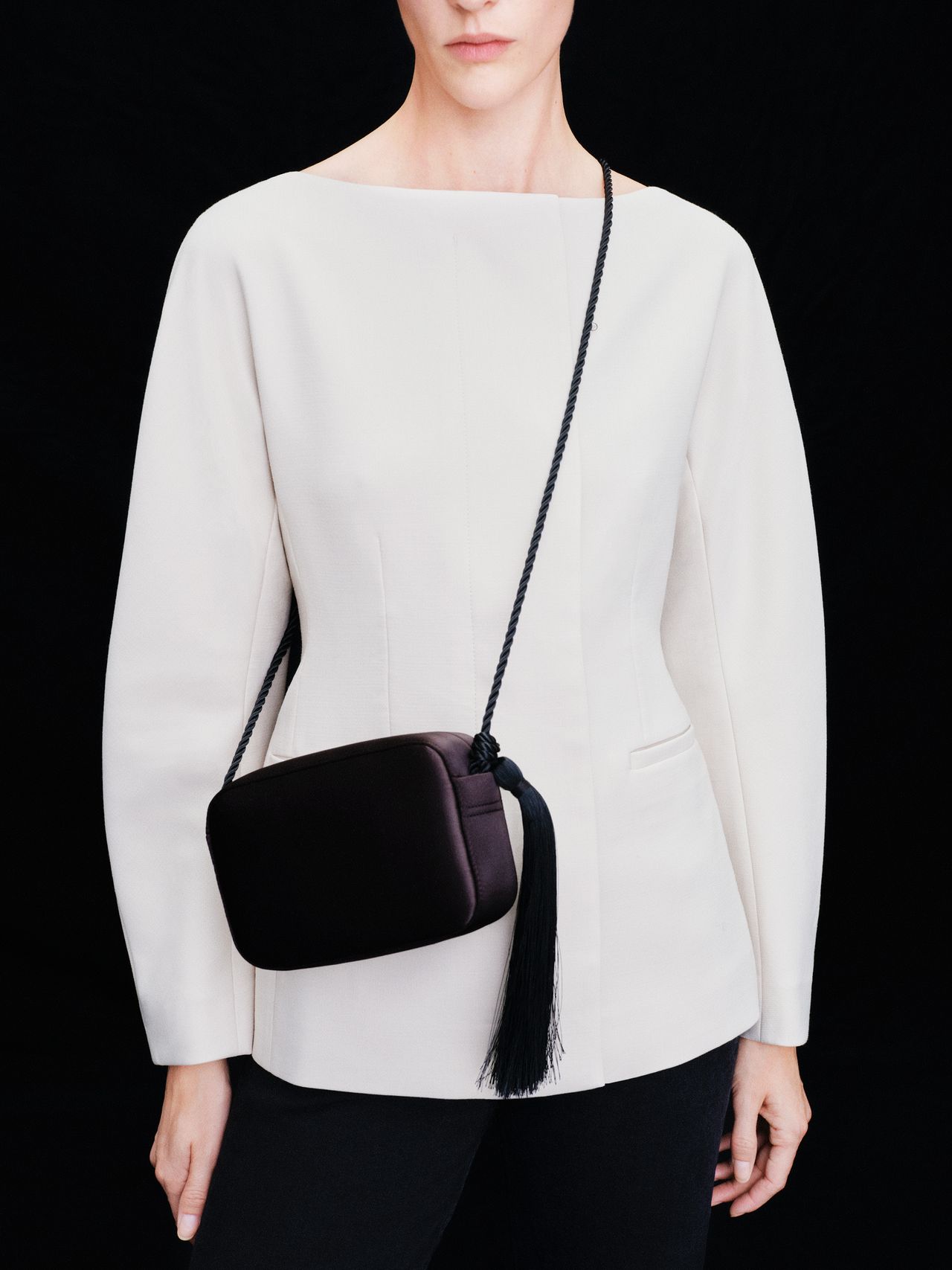 Person in a white long-sleeve top and black pants, holding a small black bag with a tassel over their shoulder, against a black background.