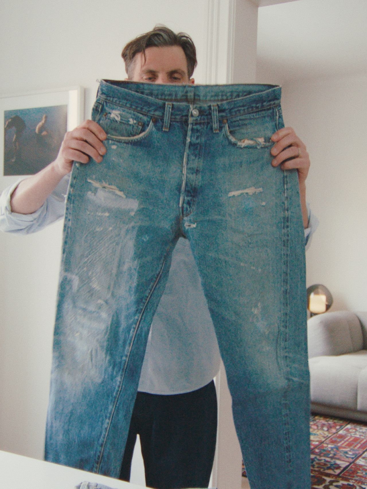 A person holding up a pair of distressed blue jeans in a modern, bright room with a couch and framed picture in the background.