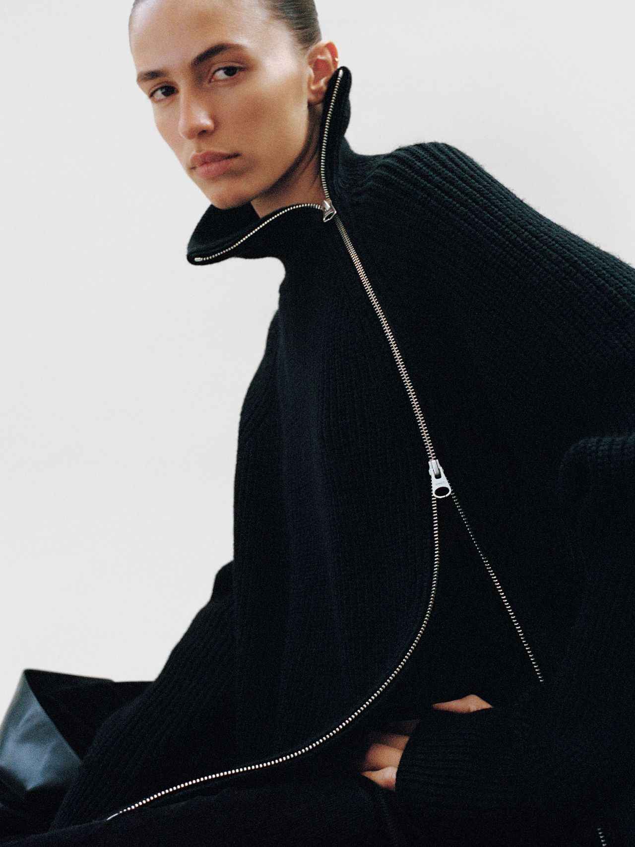 Person wearing a black, high-collared sweater with a prominent zipper, looking at the camera against a plain background.