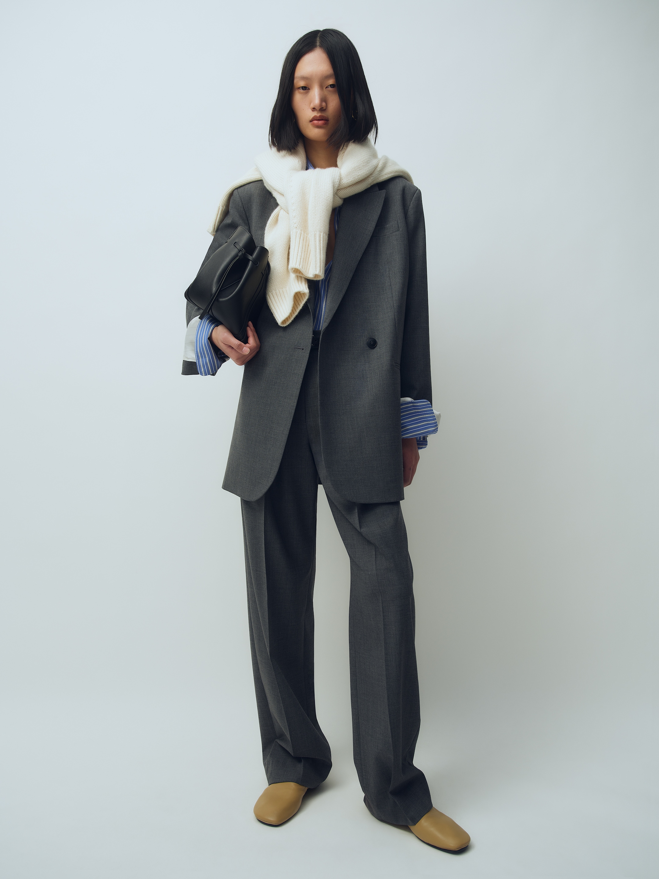 Person wearing a gray oversized suit, a blue striped shirt, beige shoes, a white scarf around the neck, and holding a black handbag.
