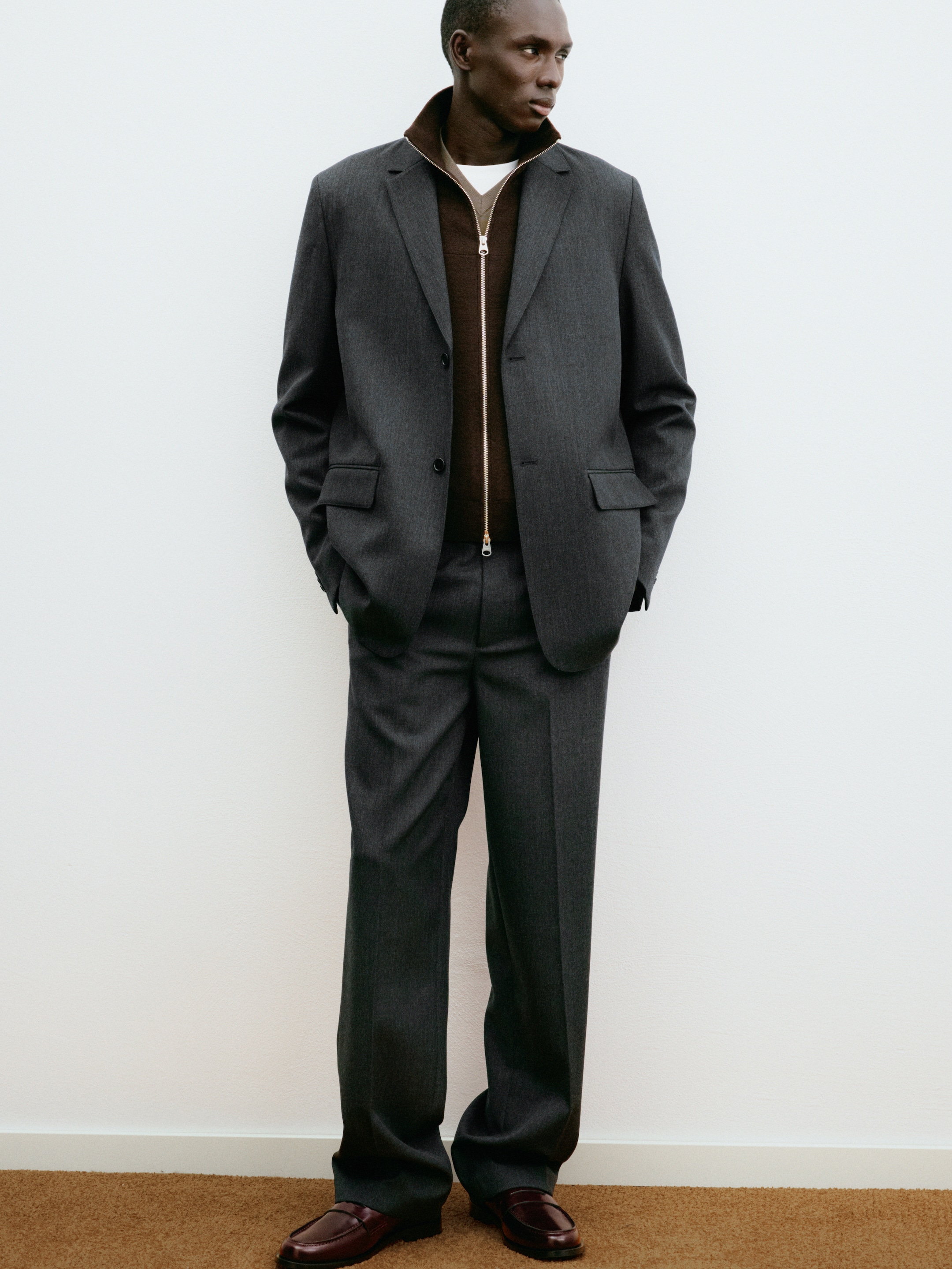 A person wearing a gray suit over a brown zip-up sweater stands with hands in pockets on a brown carpet against a white wall.