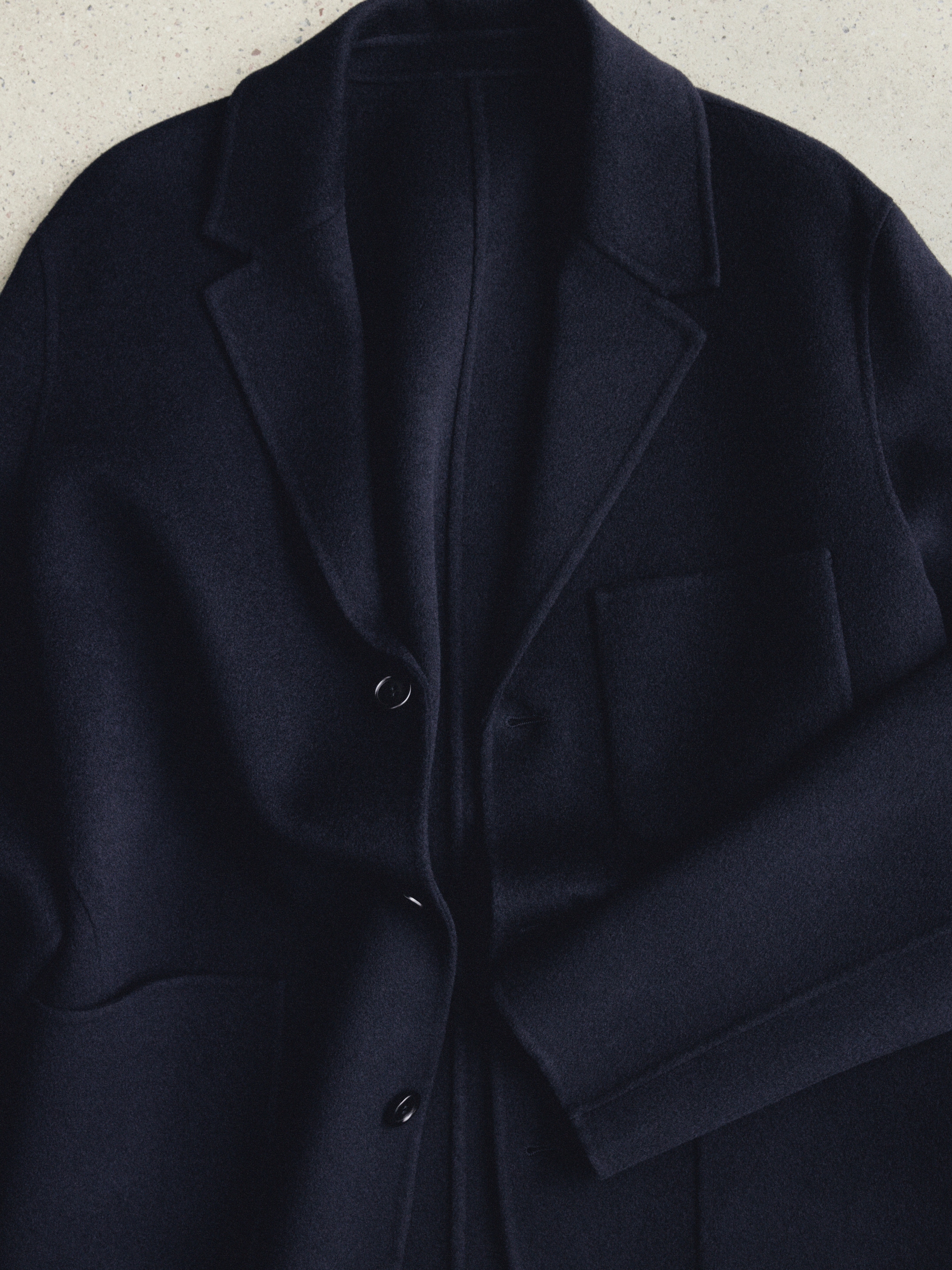 A close-up of a dark navy wool coat lying flat, featuring a notched collar, button closure, and front pocket, showcased on a light surface.