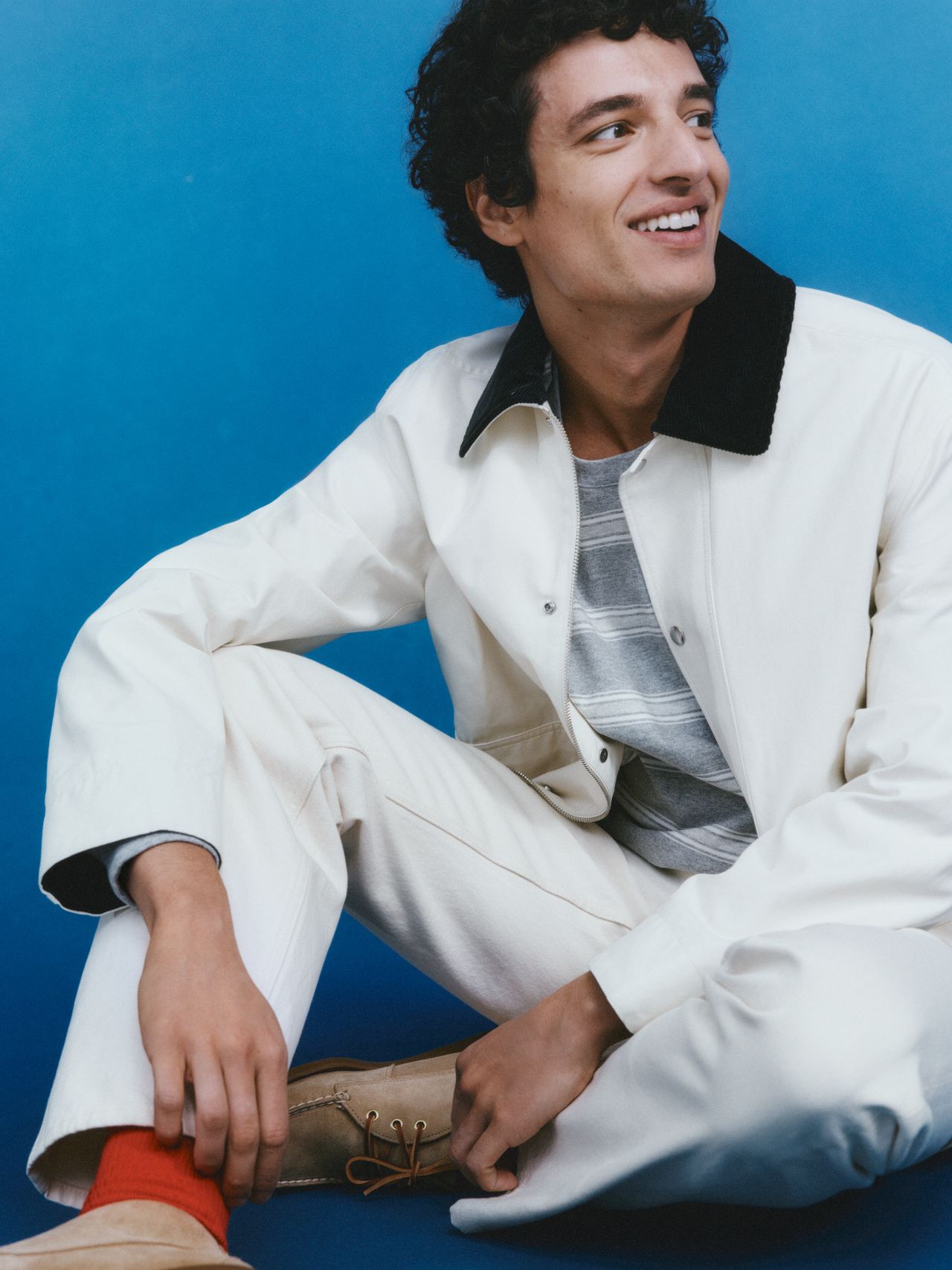 Young man in a white jacket and pants, gray striped shirt, and red socks, smiling while seated against a blue background.