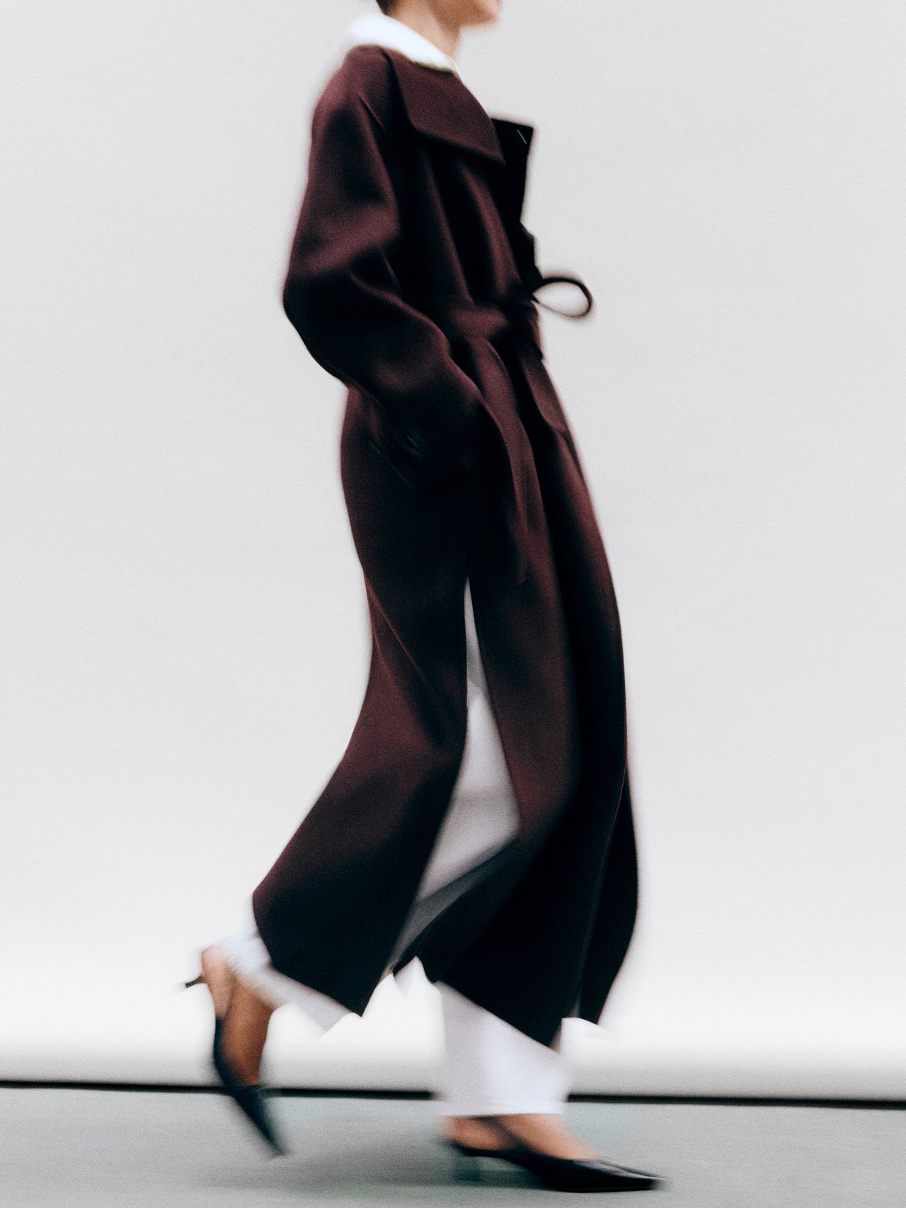 Person in a long, dark coat and white pants walking, with blurred motion, against a plain background.