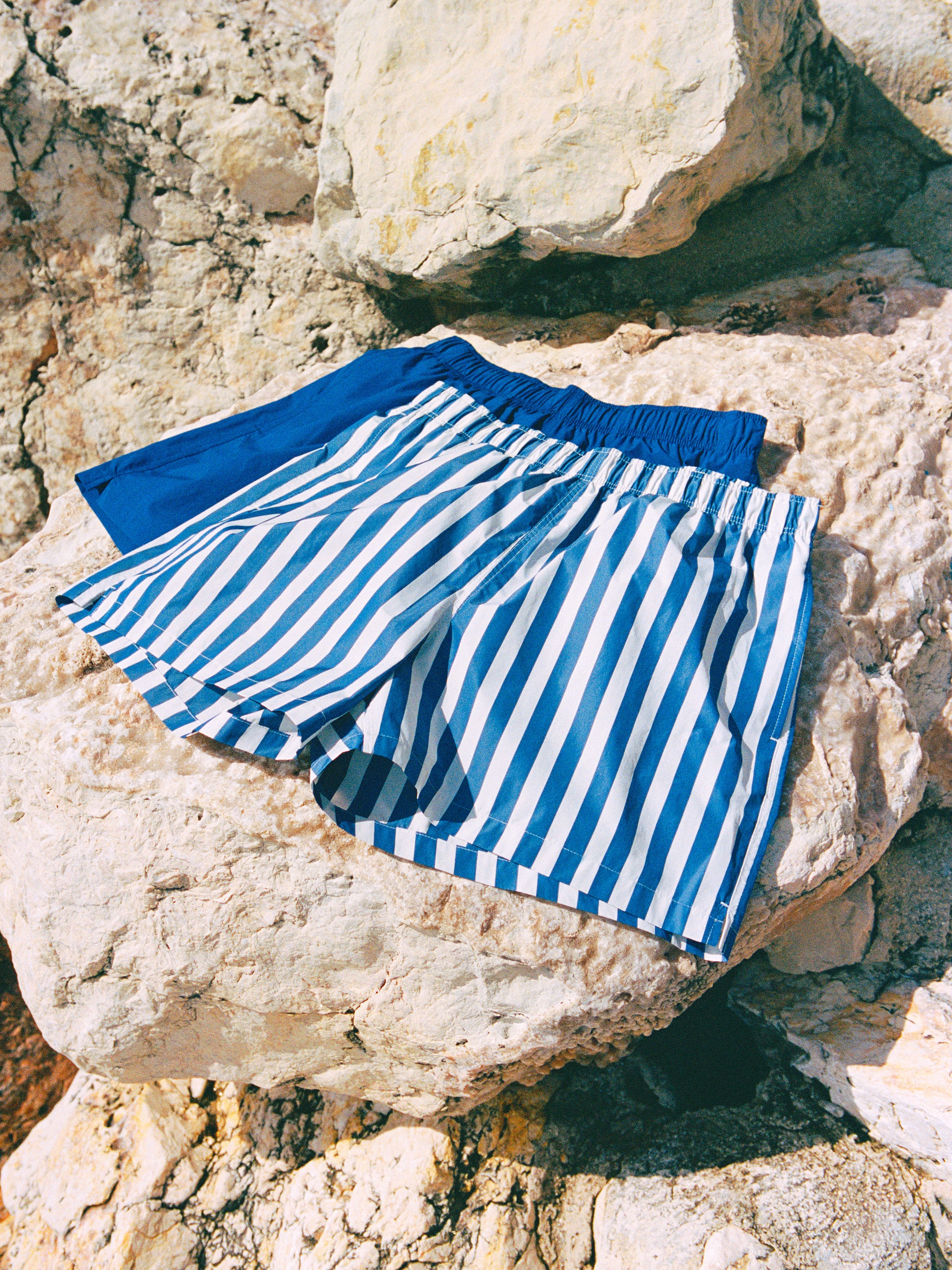 Blue and white striped shorts and solid blue shorts are laid on large, sunlit rocks.