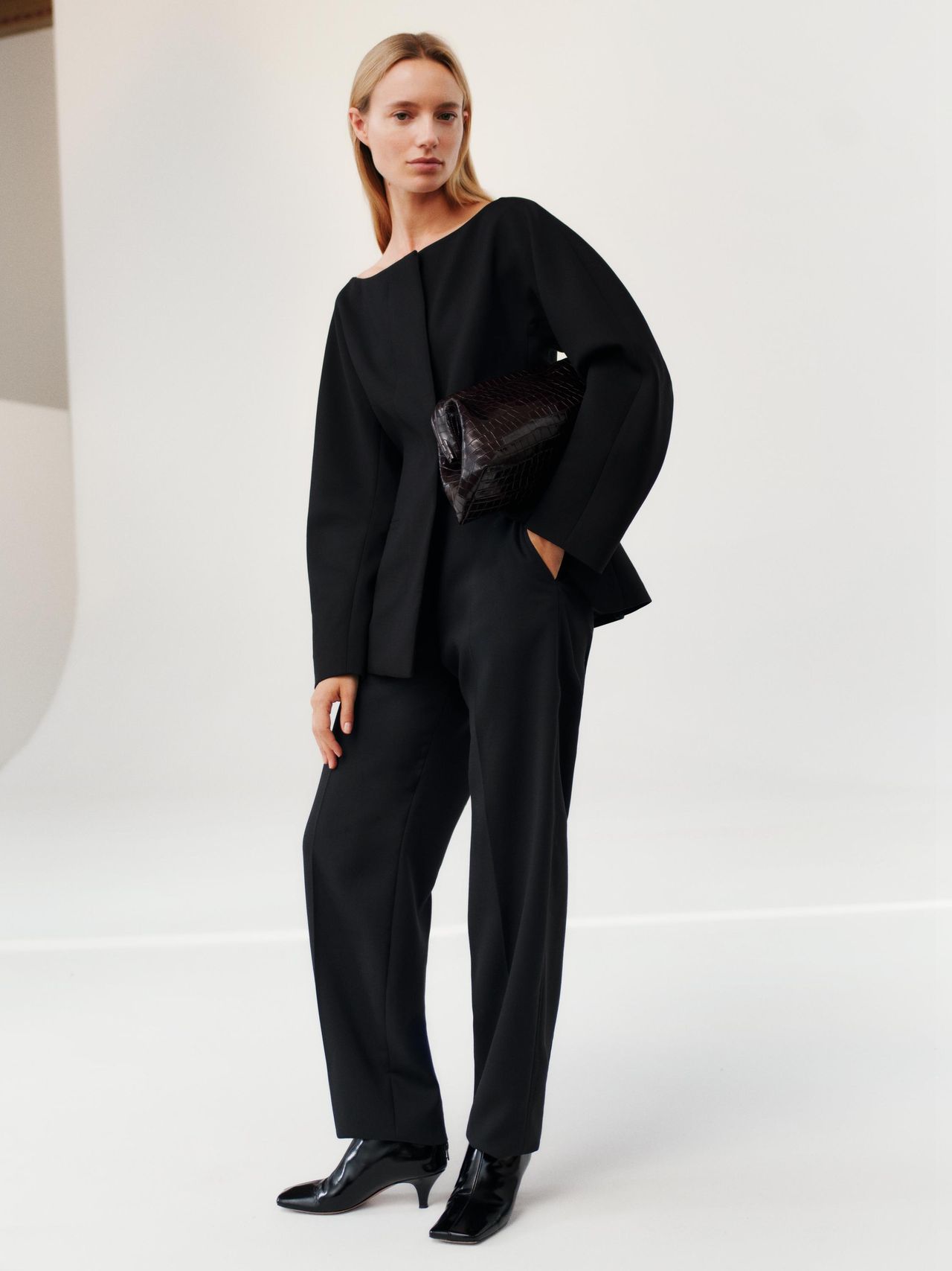 Woman in a sleek black suit and heeled boots, holding a textured black clutch, stands against a minimalist white background.