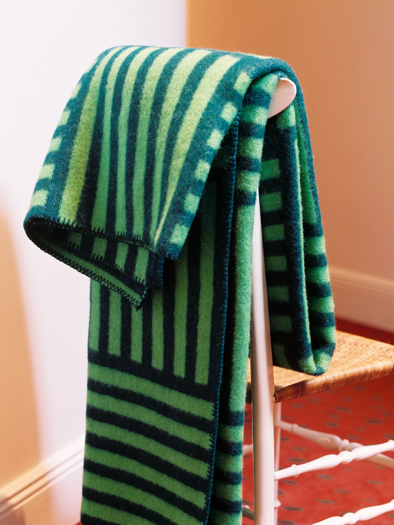 A green and black striped blanket draped over a white chair with a woven seat, set against a peach wall and red carpet.
