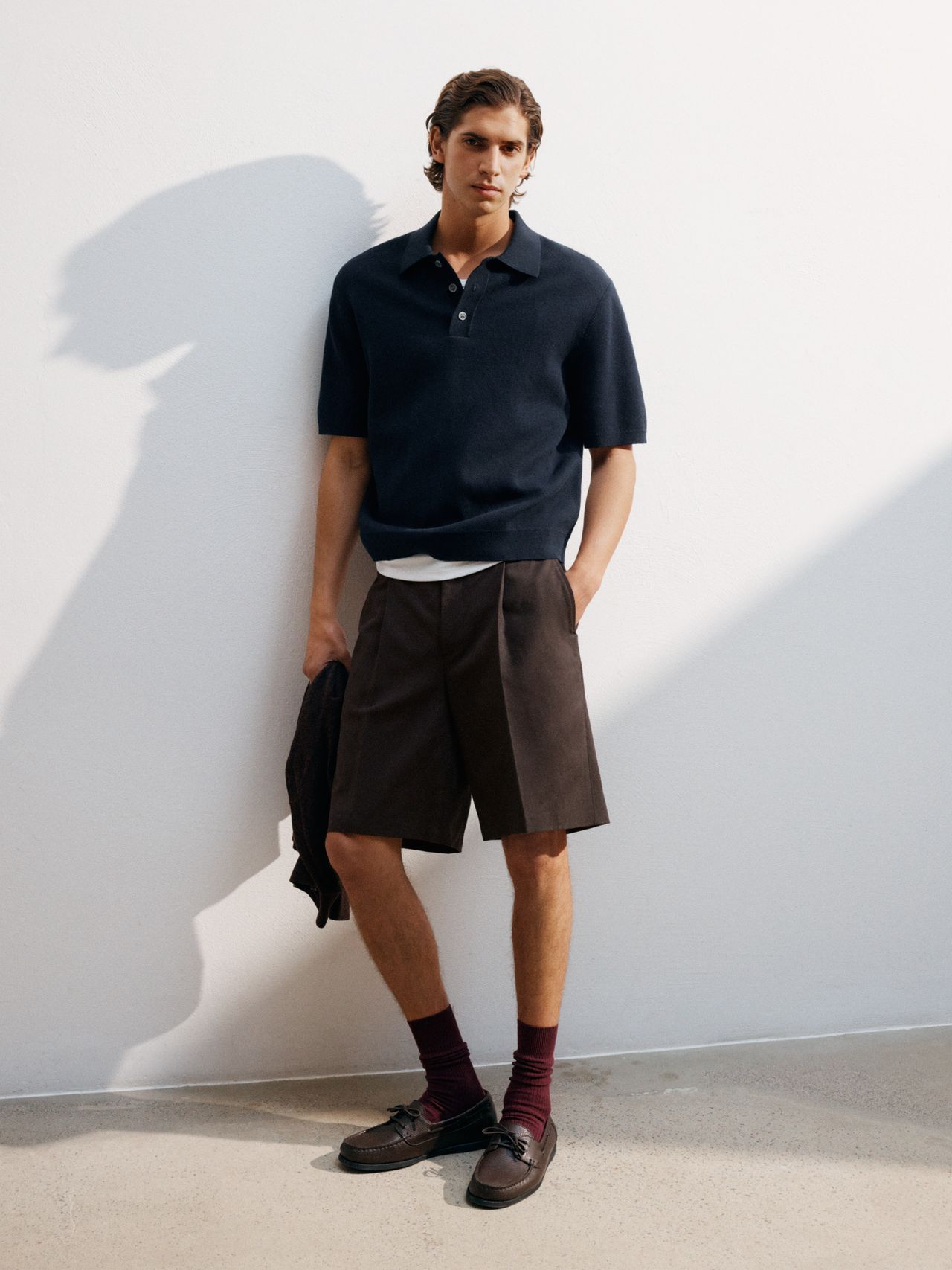 A man in a navy polo shirt and brown shorts leans against a white wall, holding a jacket. He is wearing maroon socks and brown loafers.