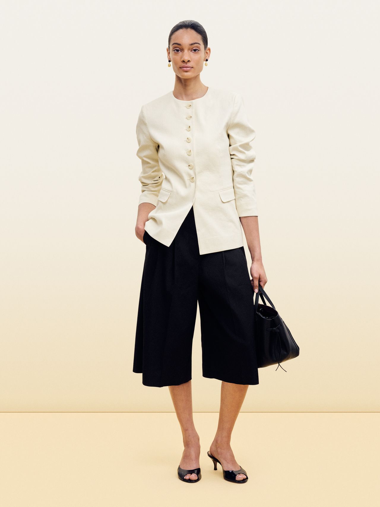 Person wearing a cream jacket, black wide-leg pants, and black heels, holding a black handbag against a beige background.