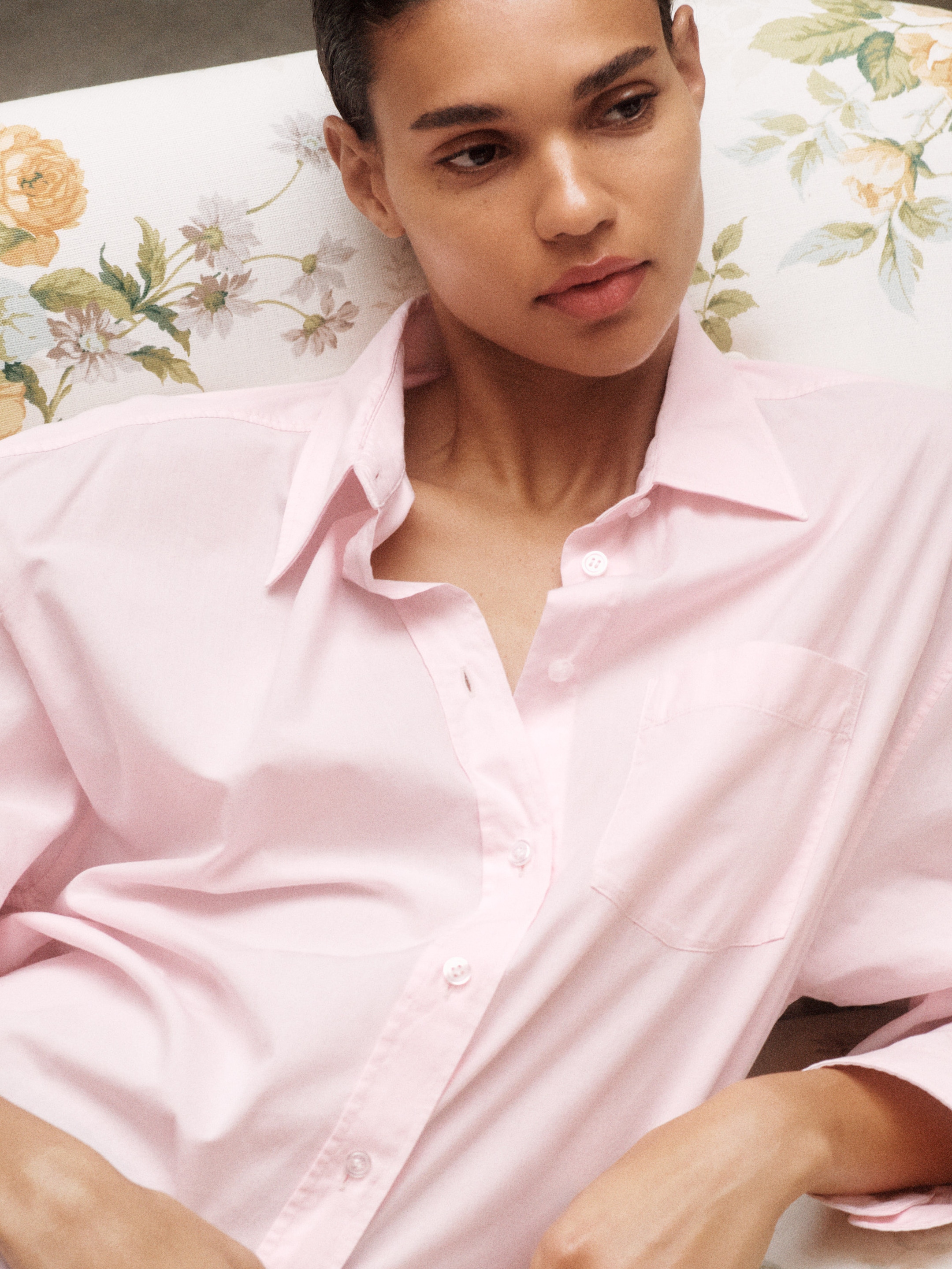 Person in a soft pink shirt, reclining on a floral-patterned couch, looking thoughtful and relaxed.