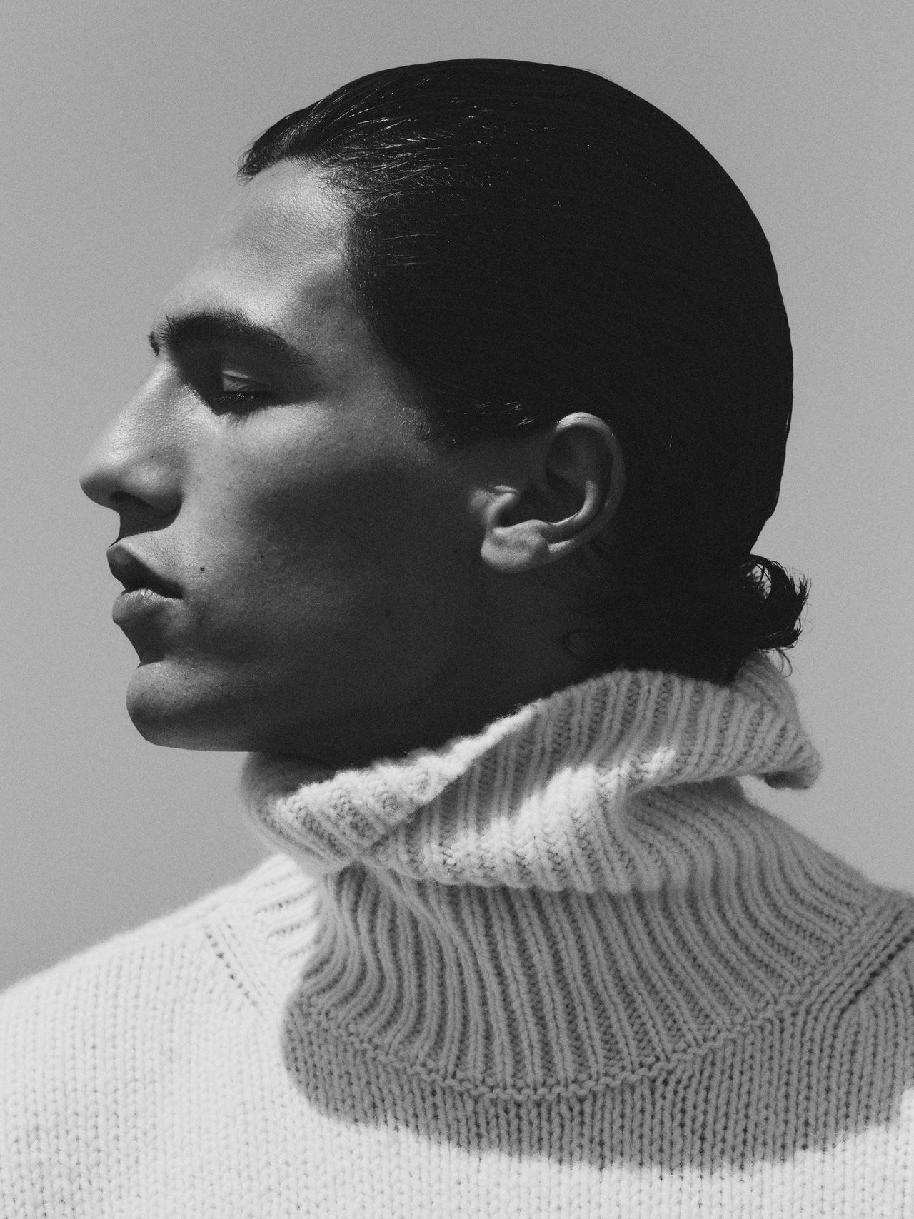 Black and white profile portrait of a person with slicked-back hair, wearing a thick, ribbed turtleneck sweater.
