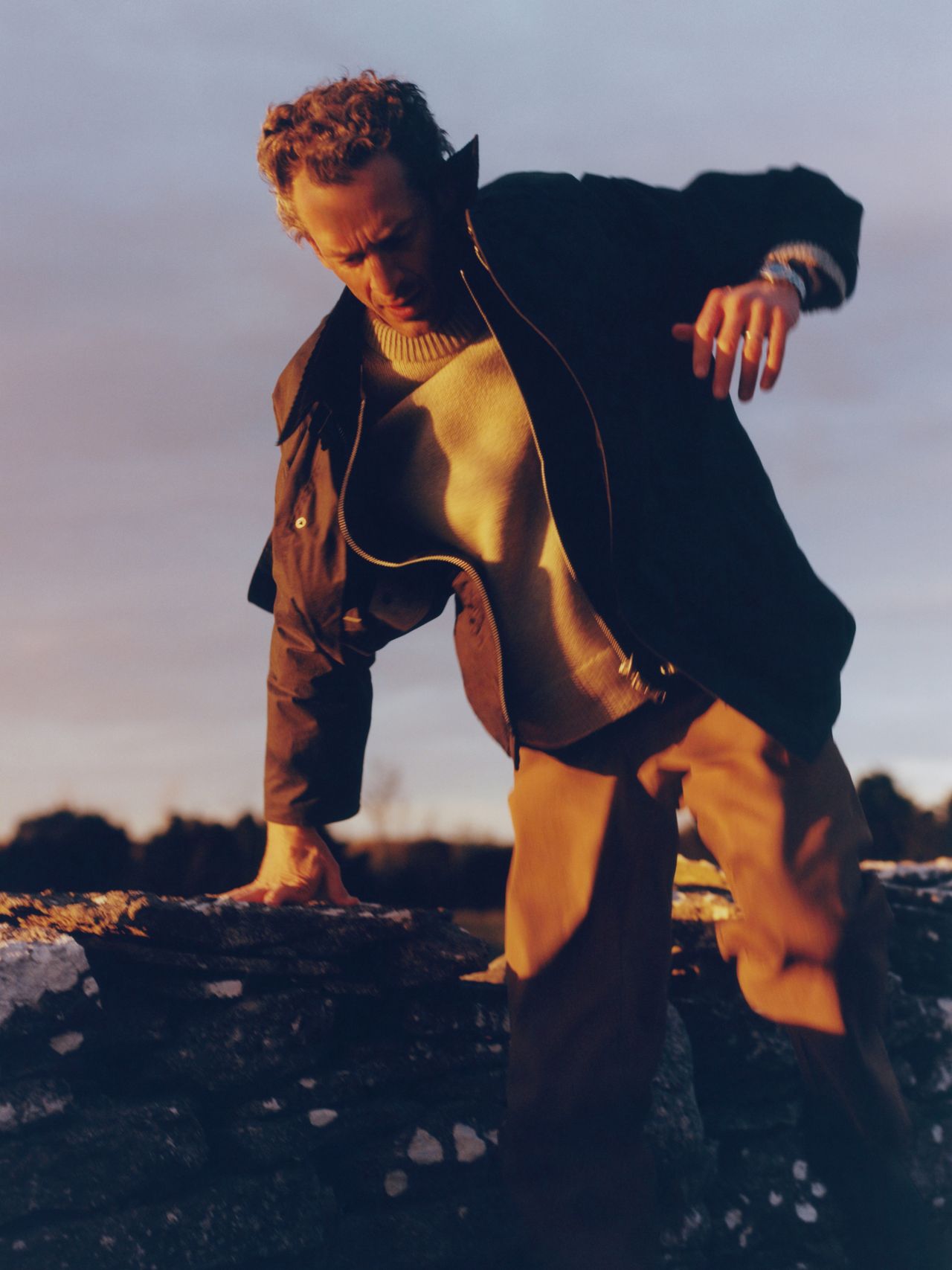 A person in a jacket and sweater climbs over a stone wall at sunset, with a blurred background of trees and a cloudy sky.