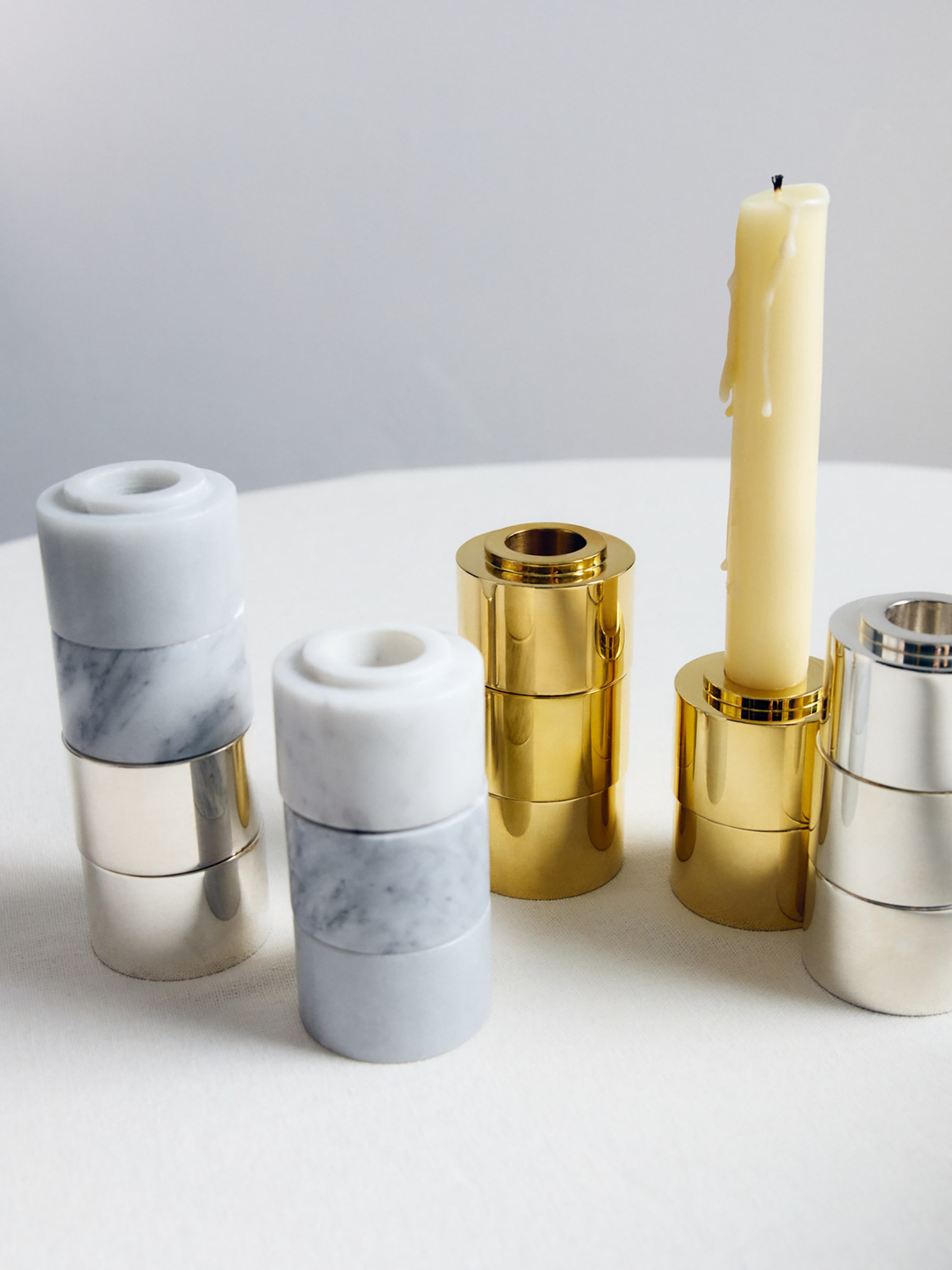 Elegant stacked candle holders in marble, gold, and silver tones are arranged on a table, with a lit yellow candle in one holder.