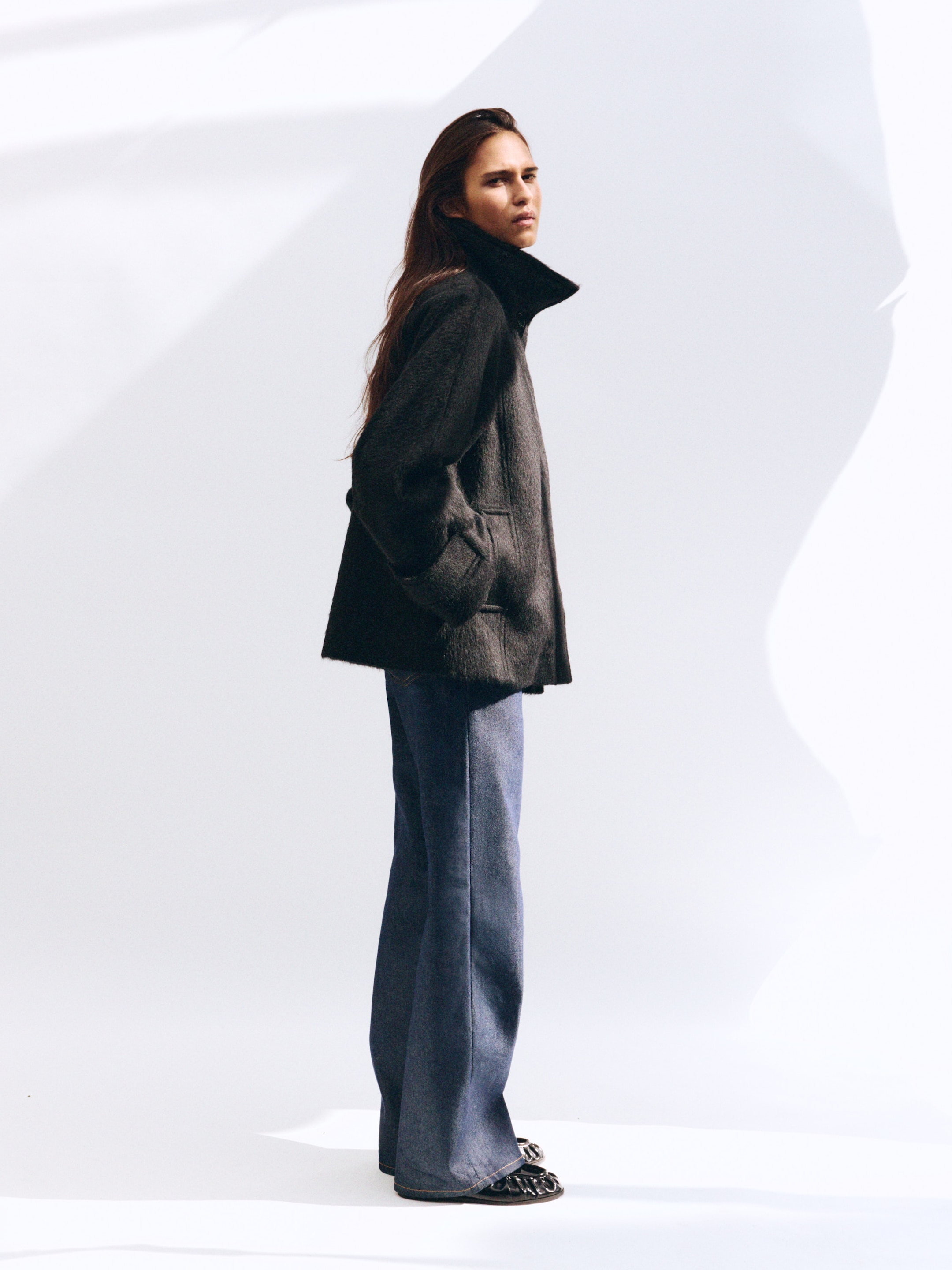 A person with long hair wearing a dark coat and flared jeans stands with hands in pockets against a light background.