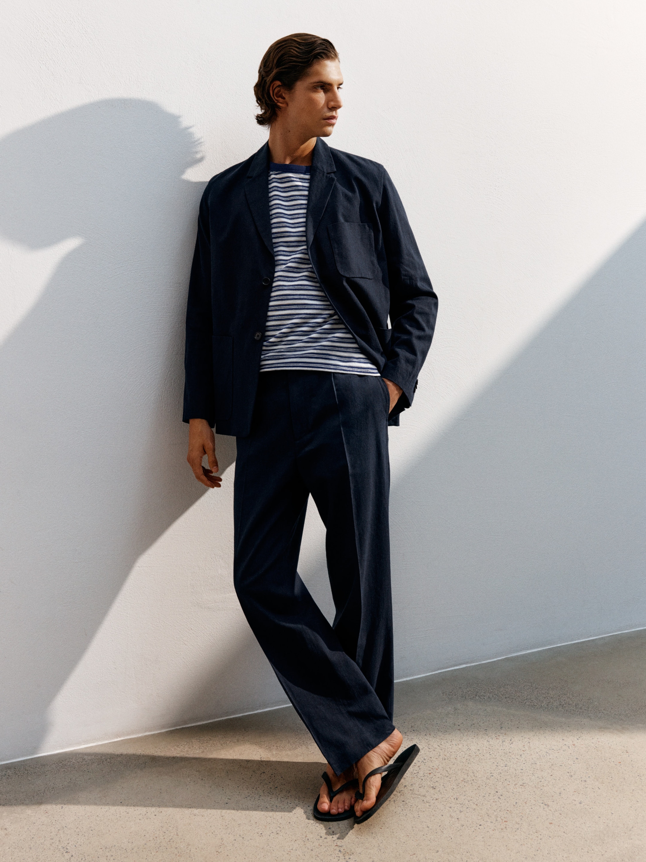 A person in a striped shirt, dark blazer, and pants leans against a white wall, wearing flip-flops in bright, natural light.