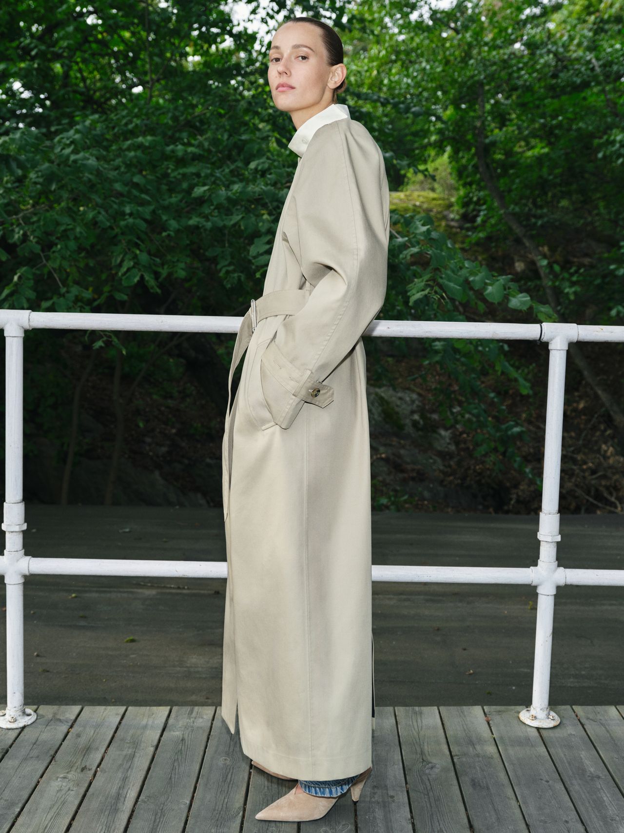 Person in a long beige trench coat and jeans stands on a wooden deck, surrounded by lush green foliage, looking sideways.
