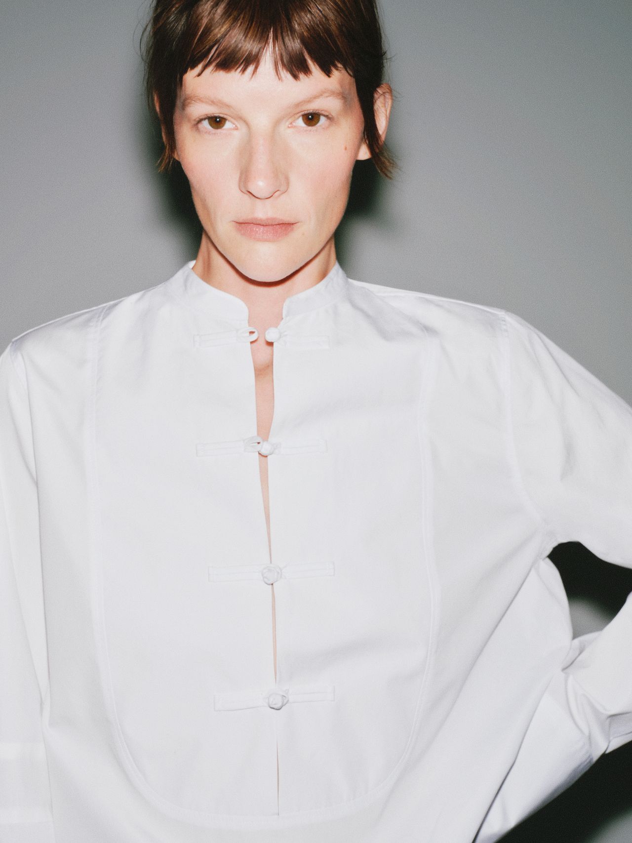 Person wearing a white, mandarin-collared shirt with button details, standing against a plain background, looking directly at the camera.