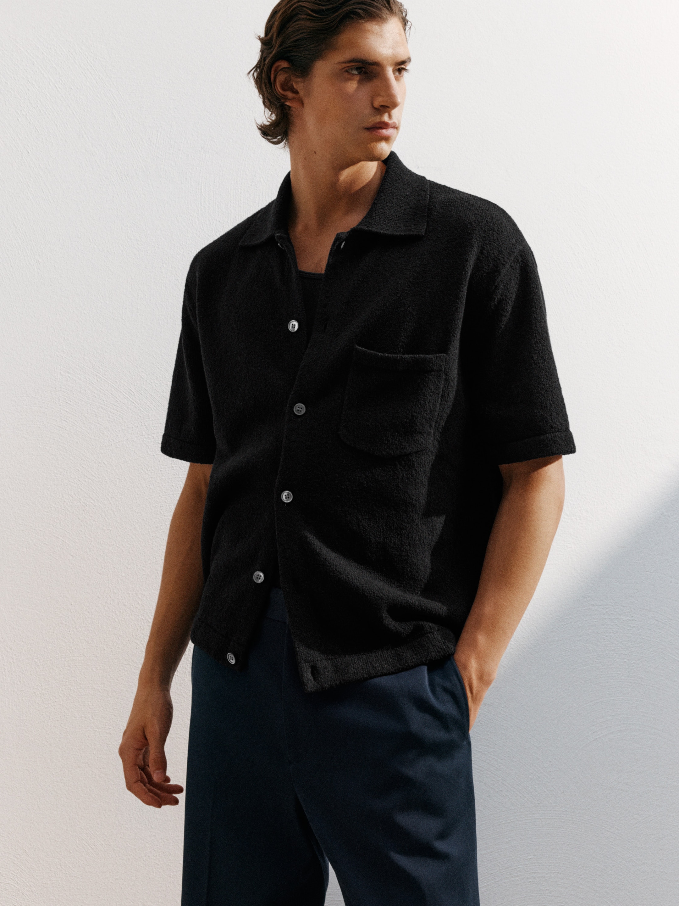 Person wearing a black button-up short-sleeve shirt and dark pants, looking to the side against a plain background.