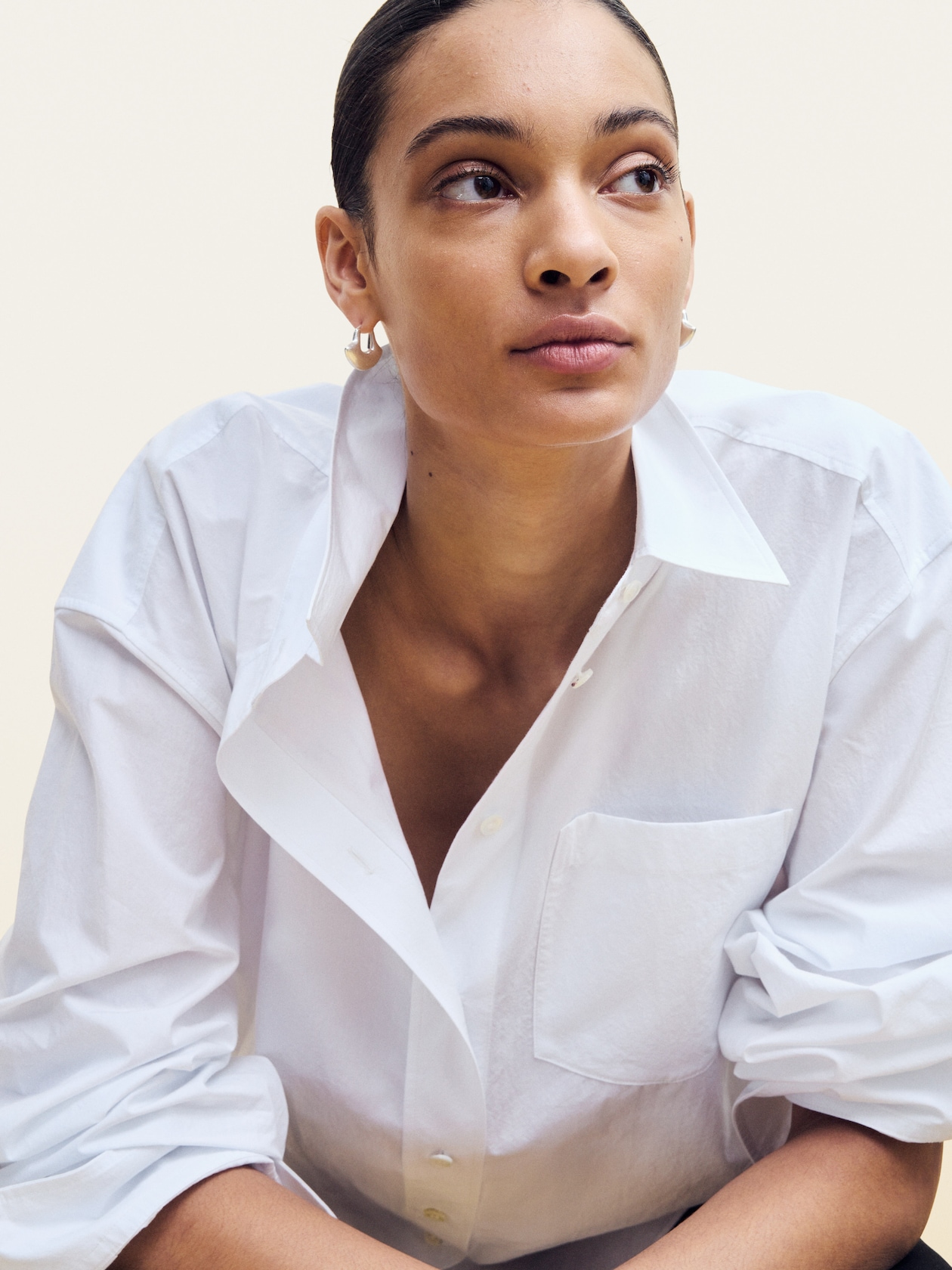 Person wearing a beige trench coat, brown sweater, white wide-leg pants, and holding a black clutch against a light background.