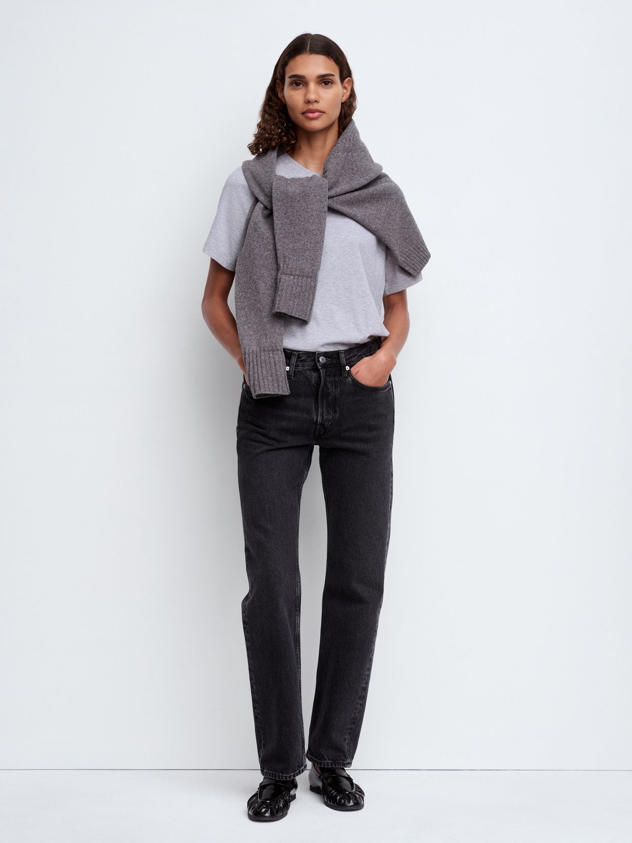 Woman in a gray t-shirt and jeans, with a sweater draped over her shoulders, stands against a white background with hands in pockets.