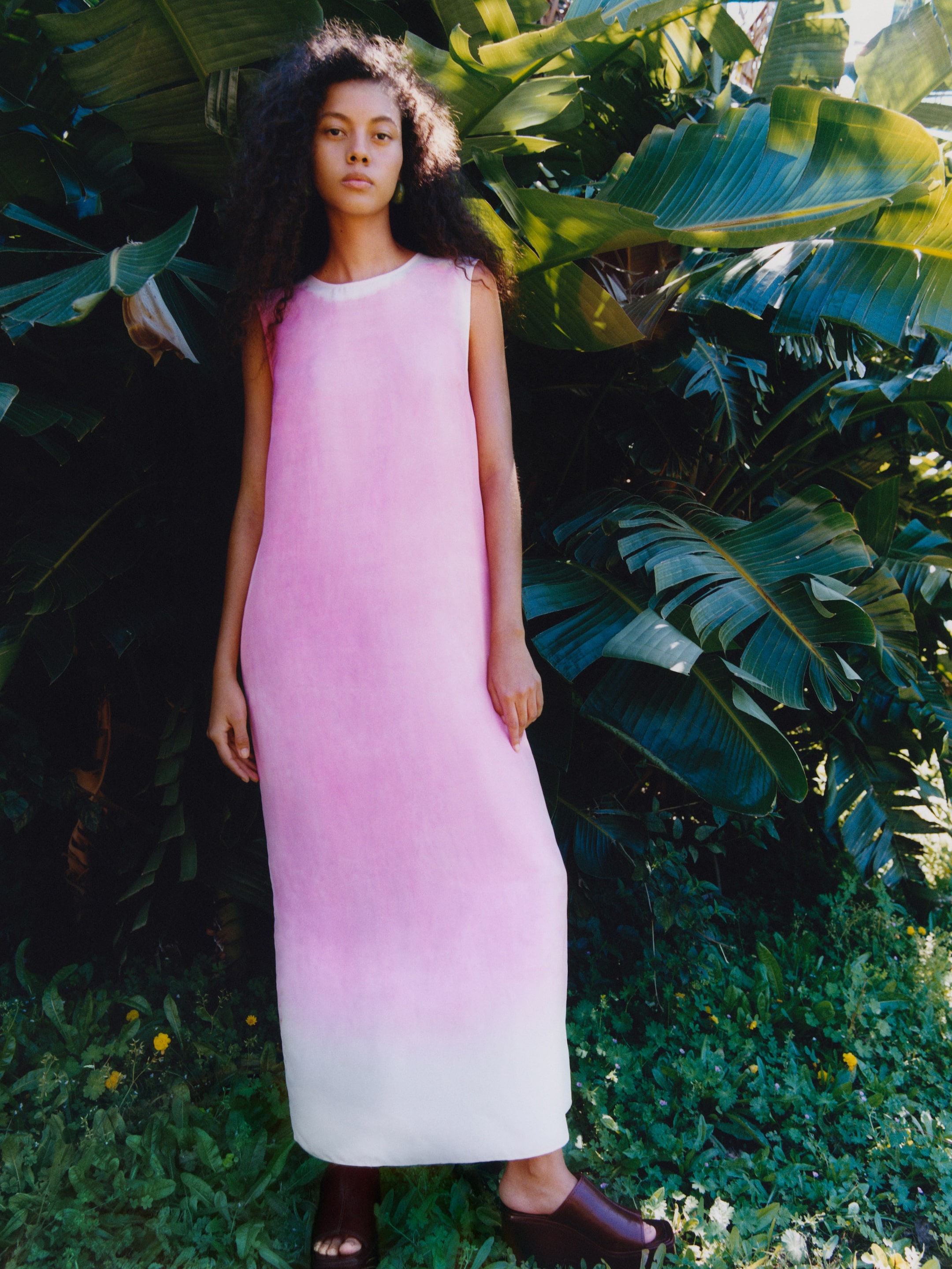 Woman in a pink and white gradient dress stands in front of lush green foliage, with sunlight filtering through the leaves.