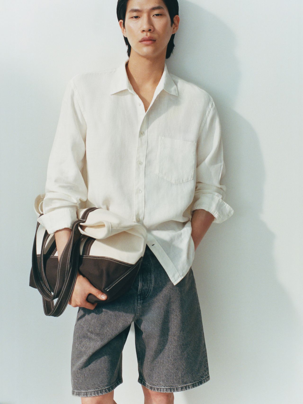 Person in a white shirt and gray shorts holds a stylish bag, standing against a light background.