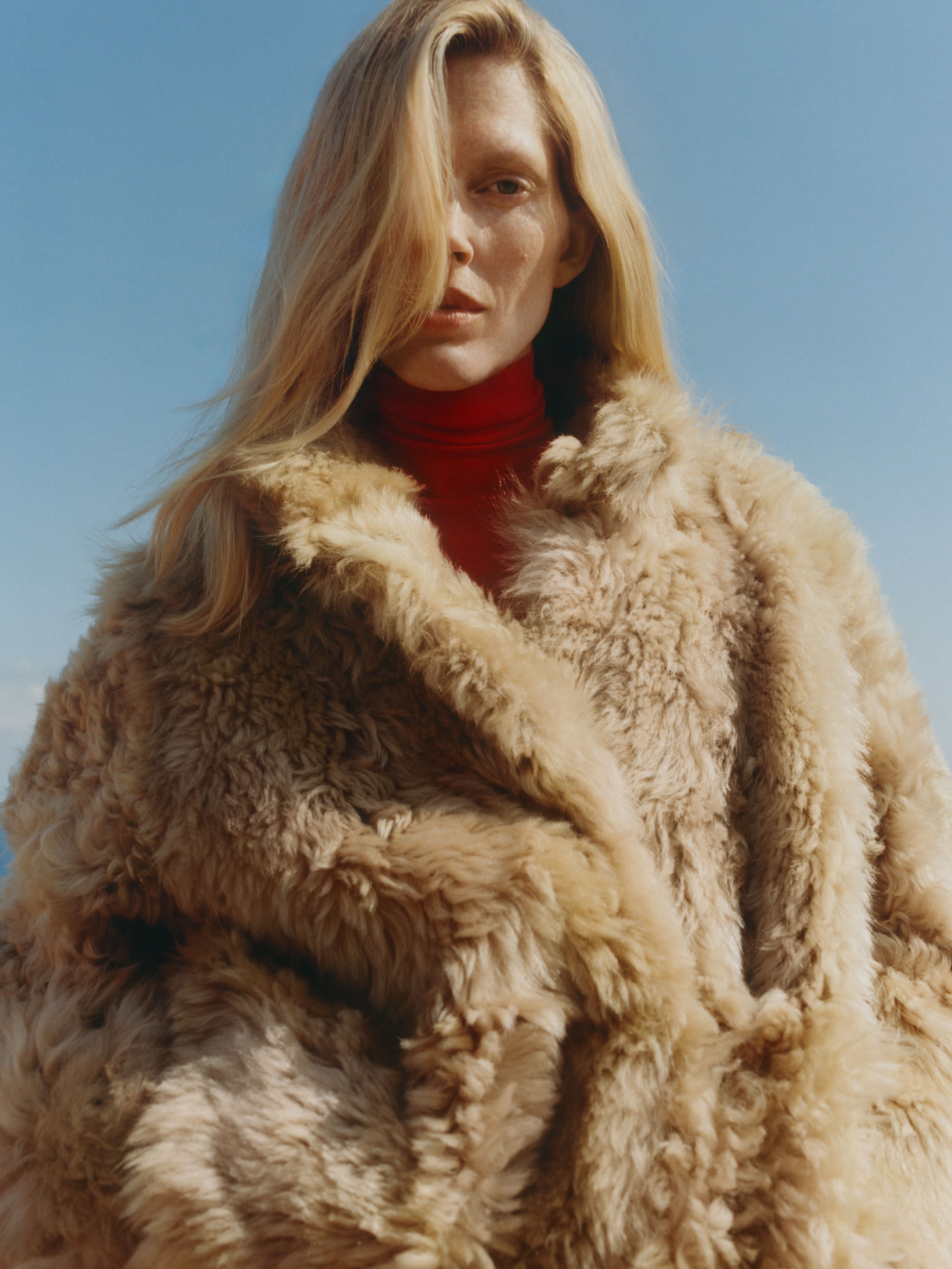 A person with long blonde hair wears a fluffy beige coat and a red turtleneck, set against a clear blue sky.