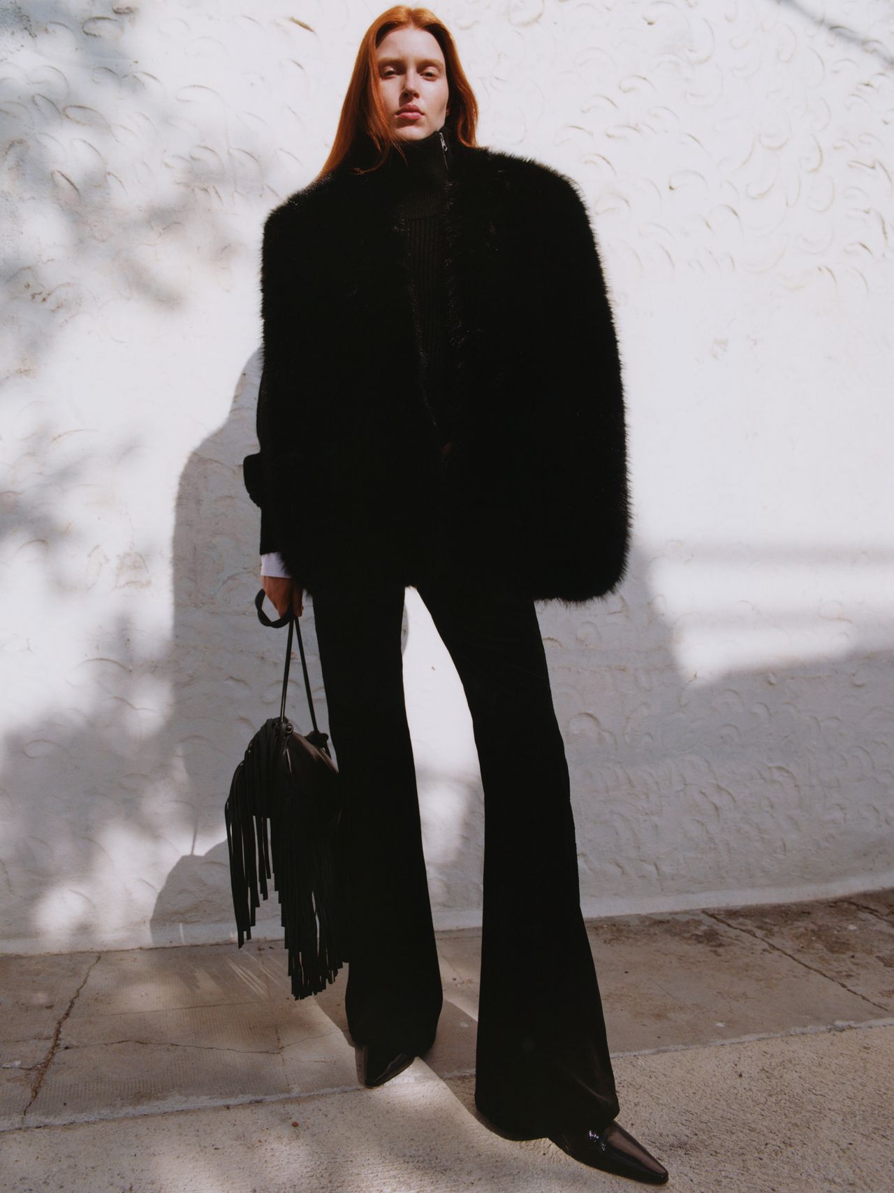 Person in black fur coat and flared pants stands against a textured white wall, holding a fringed bag. Shadows cast on the ground.