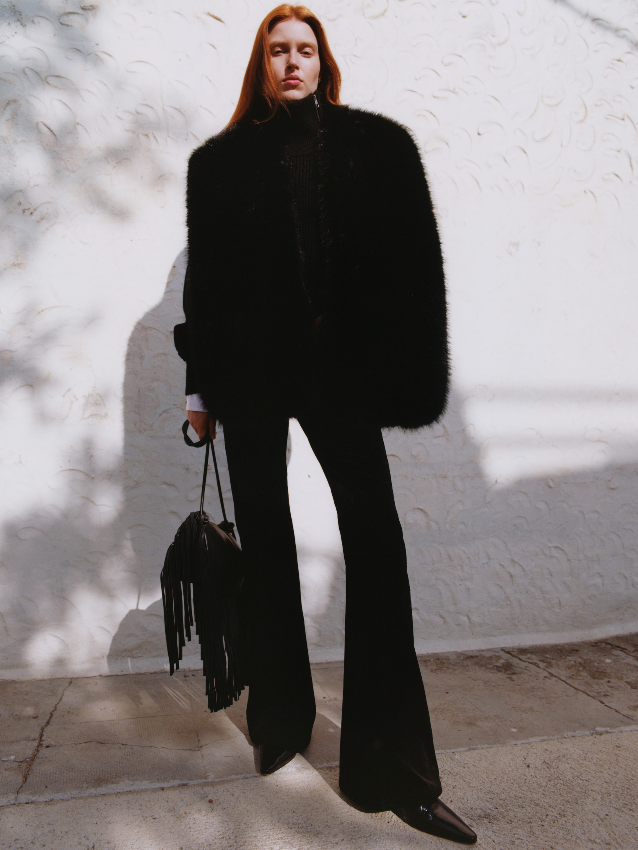 Person in black fur coat and flared pants stands against a textured white wall, holding a fringed bag. Shadows cast on the ground.