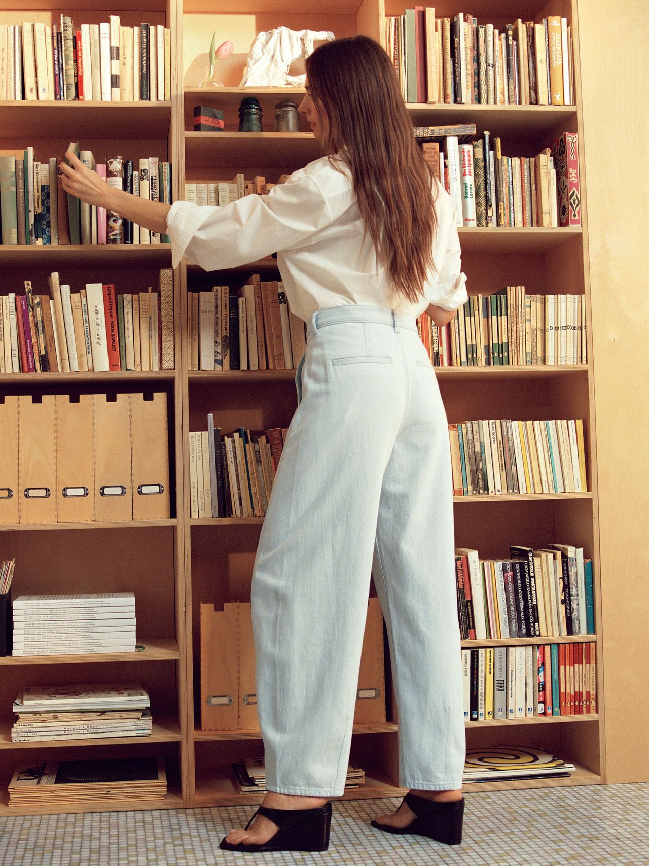 A woman with long hair wearing a white shirt and light jeans reaches for a book on a wooden bookshelf filled with books and files.