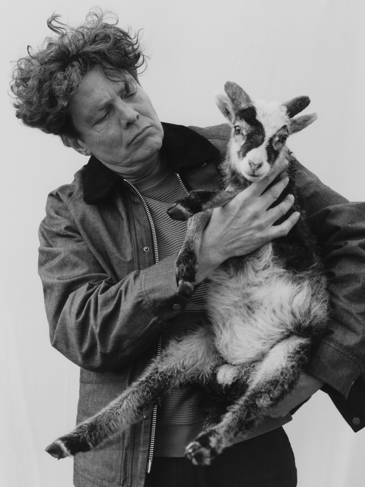Person with curly hair holding a small goat, looking at it with a curious expression. The background is plain white.
