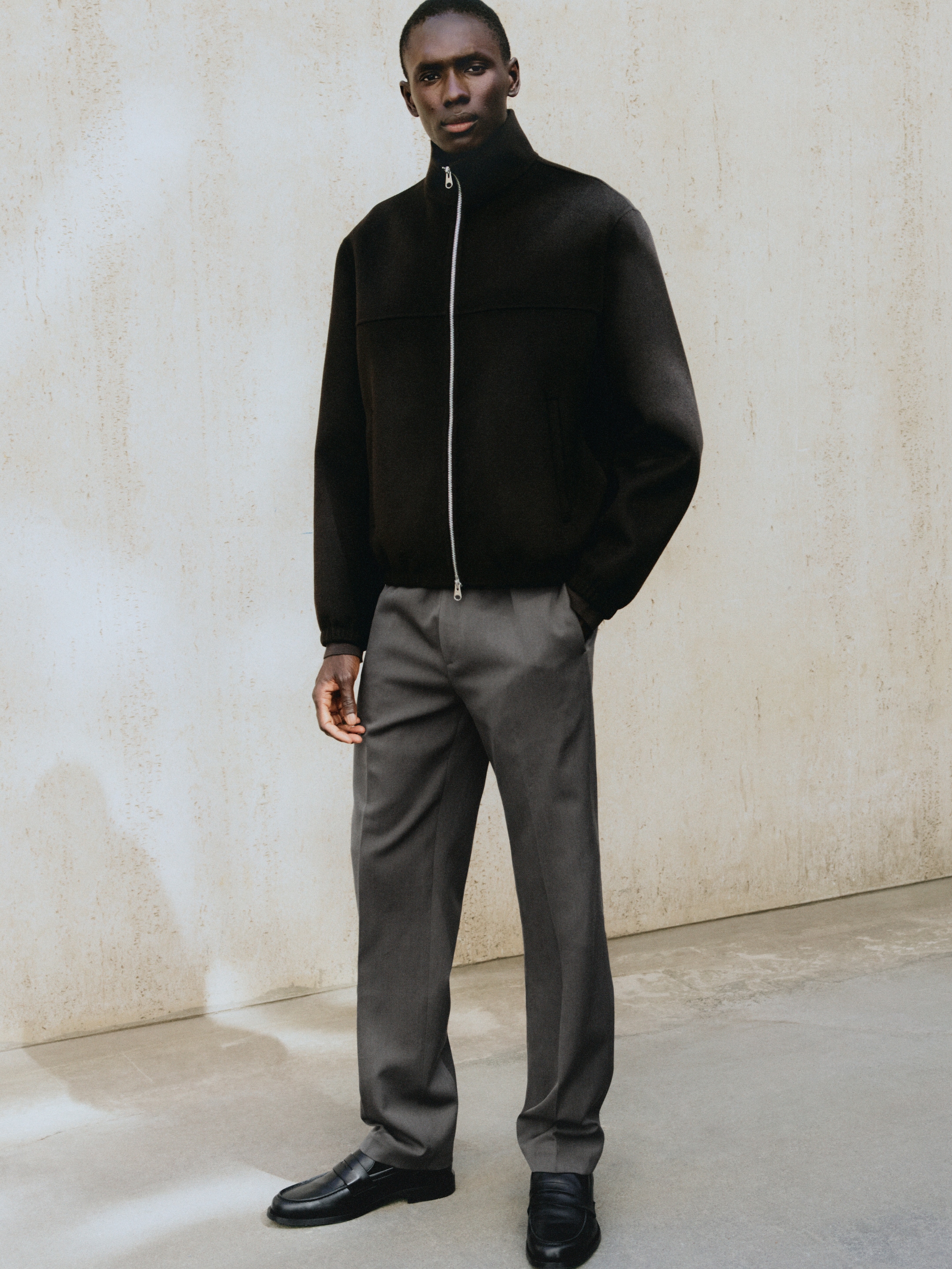 A person wearing a black zip-up jacket, gray pants, and black shoes stands against a textured neutral wall.
