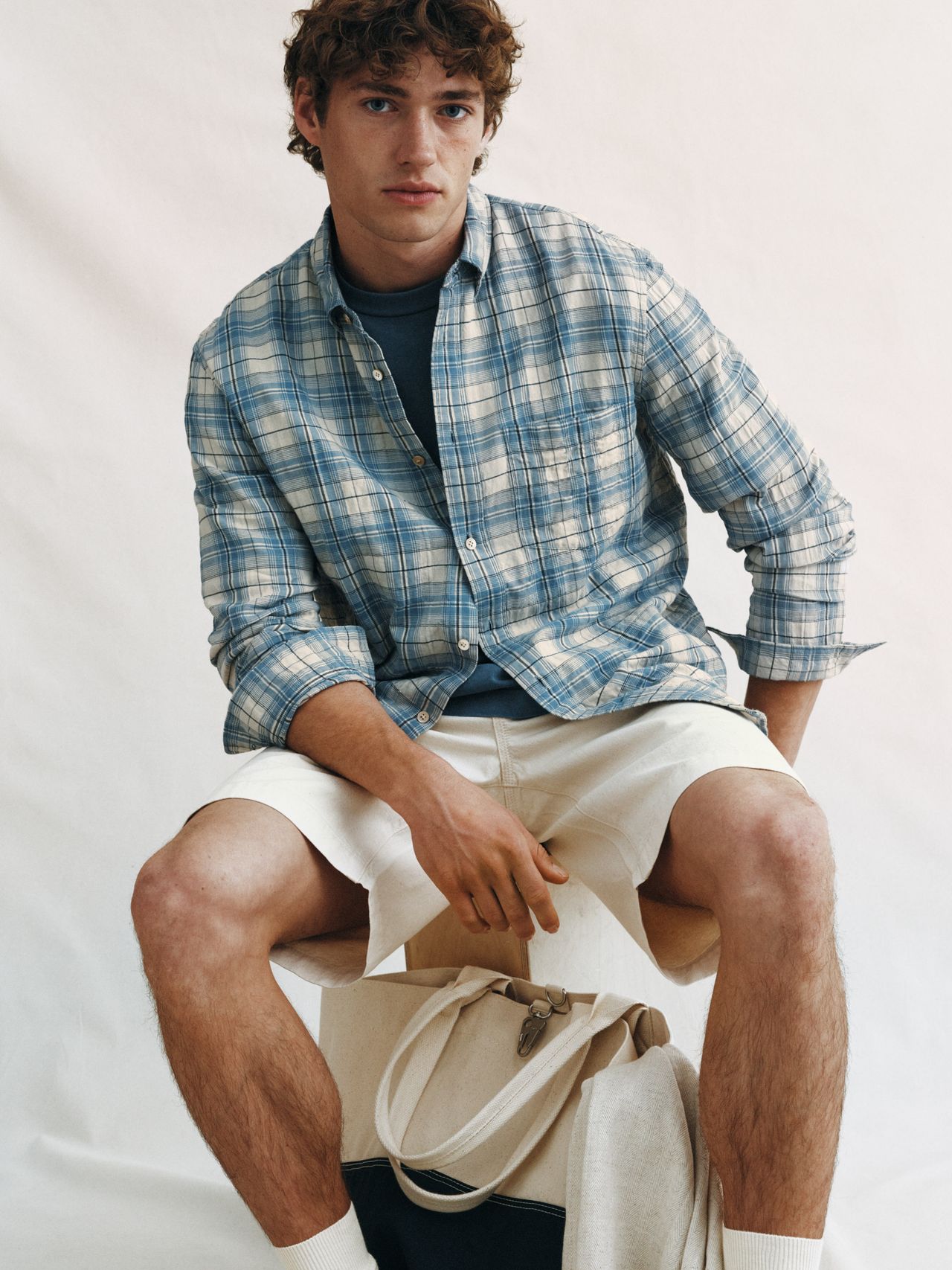 Man in a blue plaid shirt and beige shorts sits on a stool, holding a canvas tote bag, set against a light background.