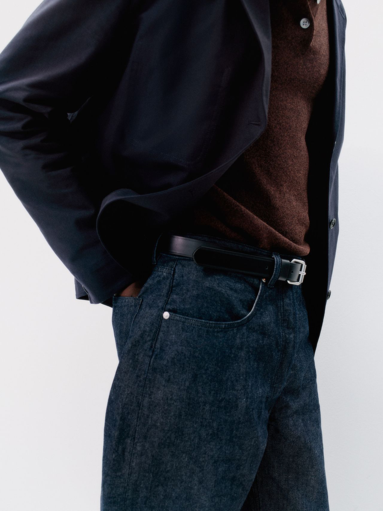 Person wearing a dark blazer, brown sweater, and blue jeans with a belt, standing against a white background.
