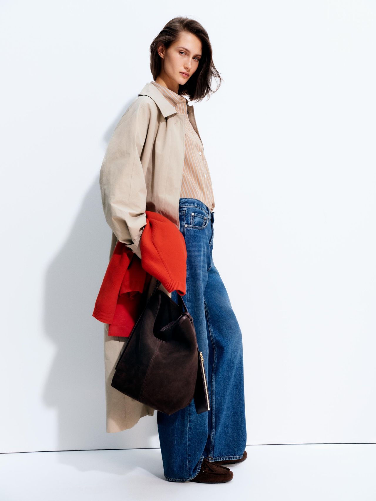 Woman in a beige coat over a striped shirt and wide-leg jeans, holding a black bag and red garment, against a white backdrop.