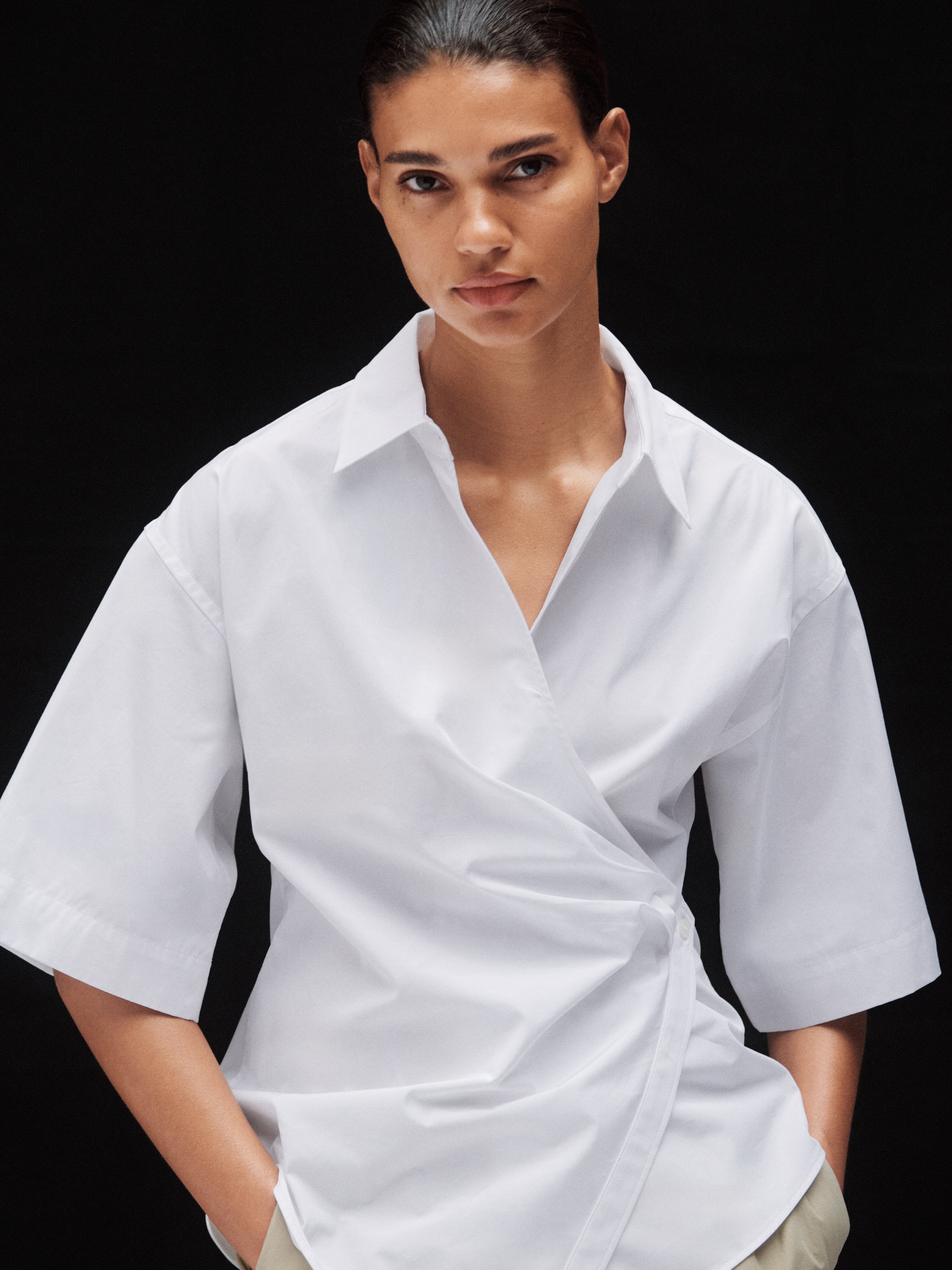 Person wearing a white wrap-style shirt with short sleeves, standing against a dark background with hands in pockets.