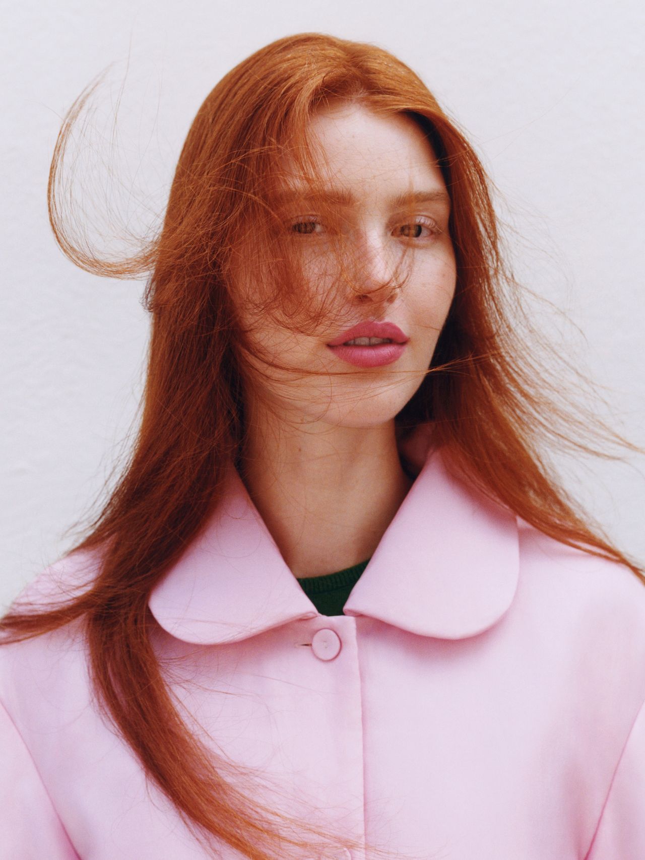 Woman with long red hair in a pink coat, standing against a white background. Her hair is gently blown by the wind.