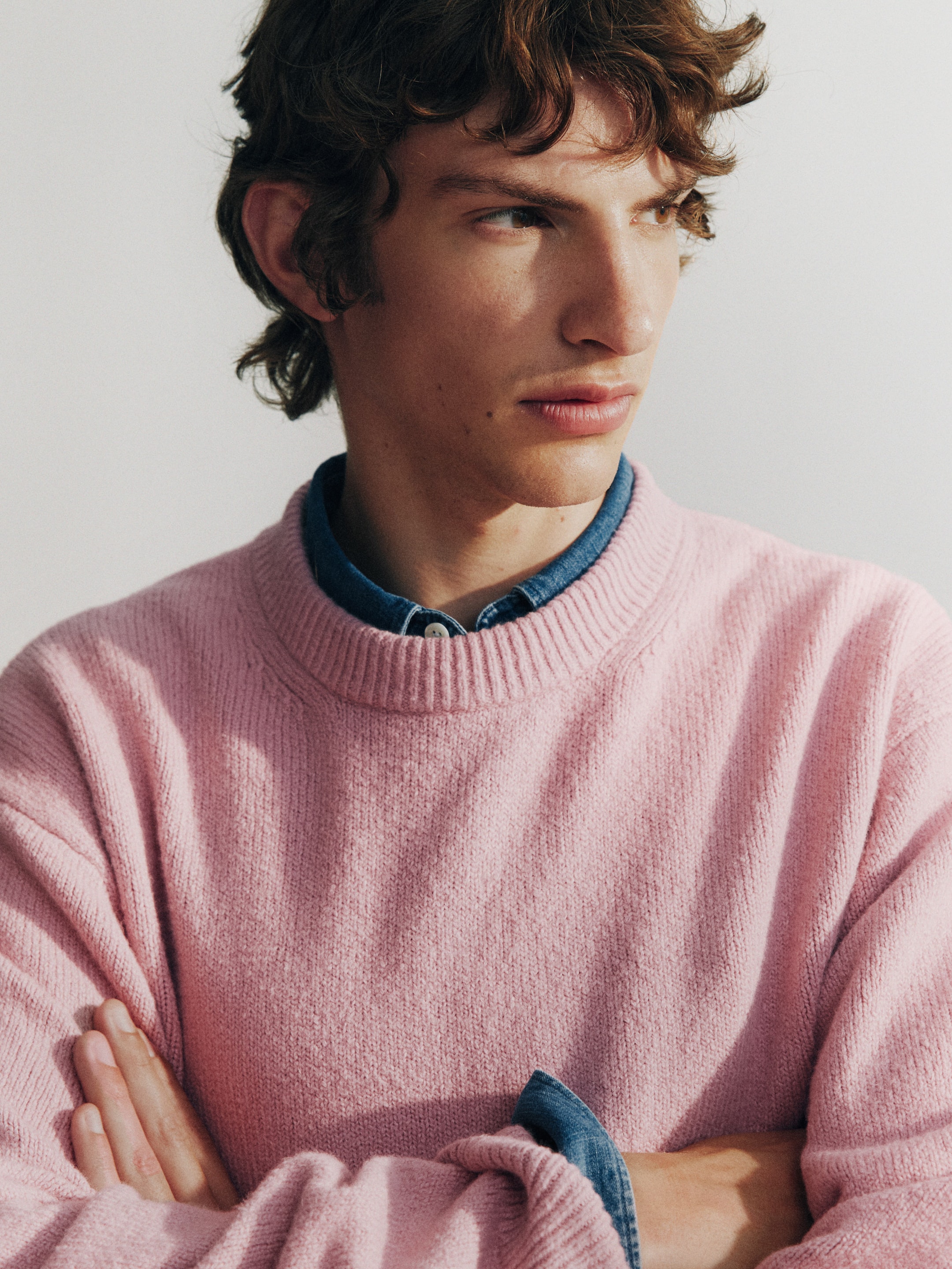 Person with curly hair wearing a pink sweater over a denim shirt, arms crossed, gazing to the side against a plain background.