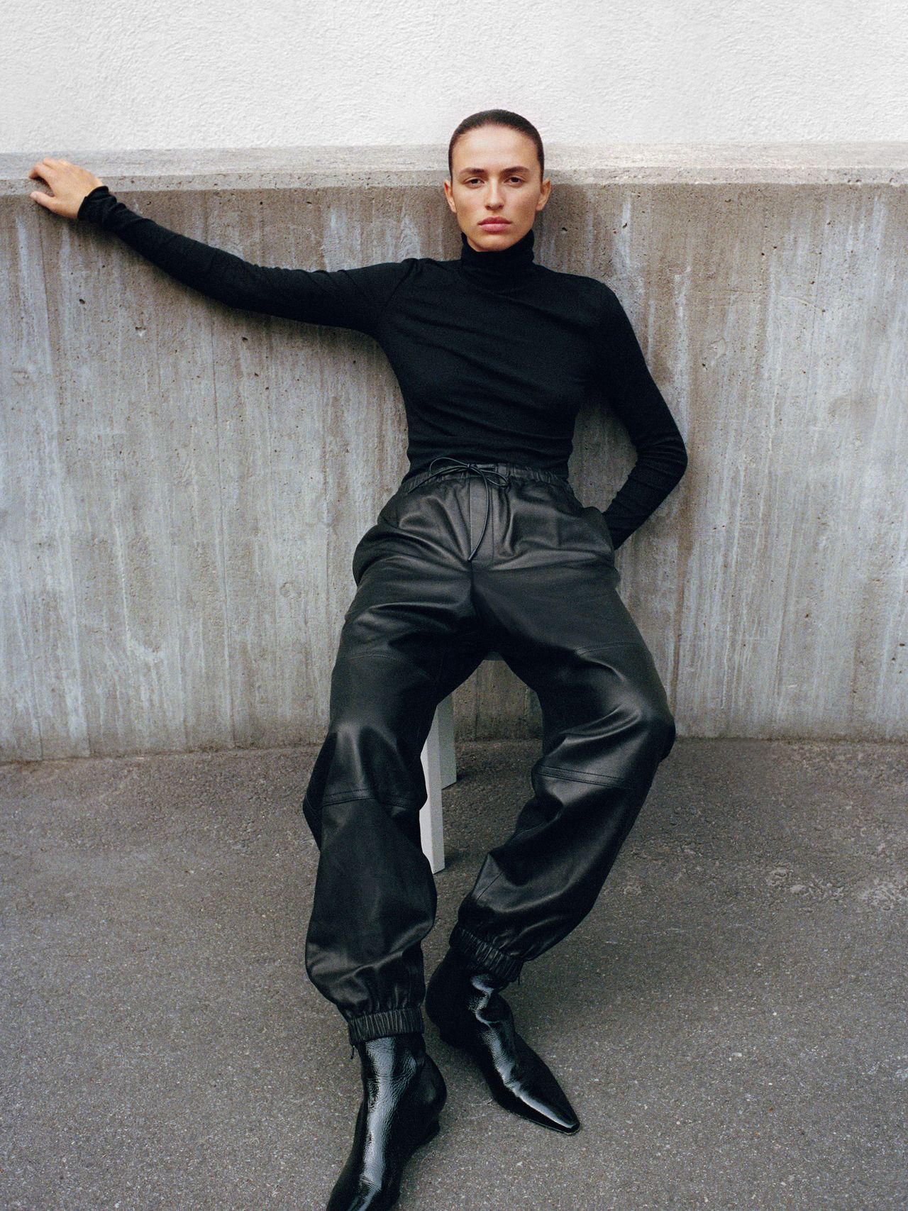 Person in a black turtleneck and leather pants sits against a concrete wall, one arm outstretched, wearing pointed black boots.