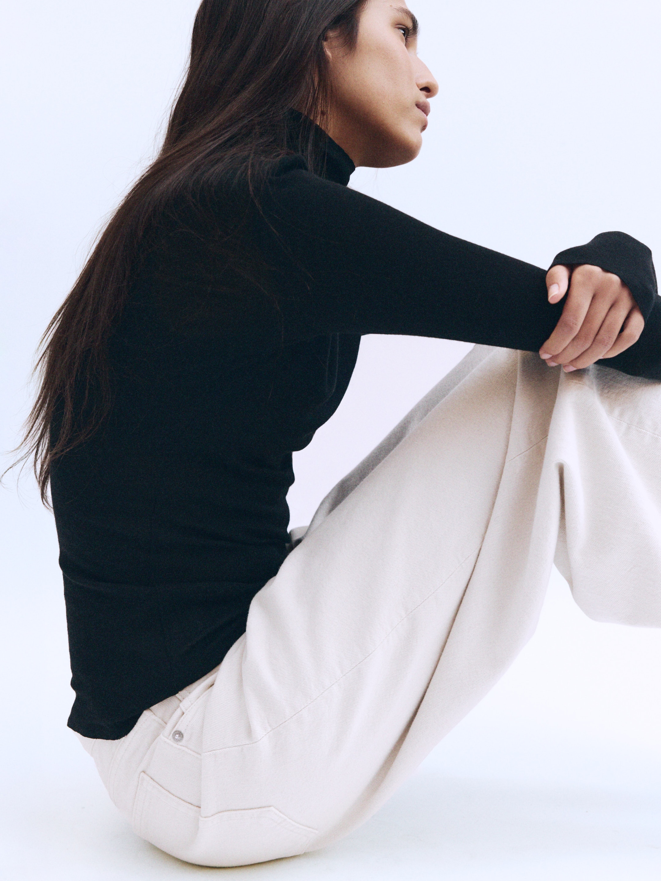 A woman in a black turtleneck and cream pants sits with knees up, gazing to the side. Her long hair flows down her back.