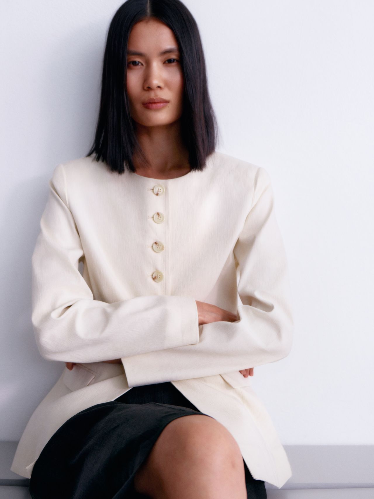 A person with straight black hair wears a cream buttoned jacket and sits with arms crossed, against a light background.