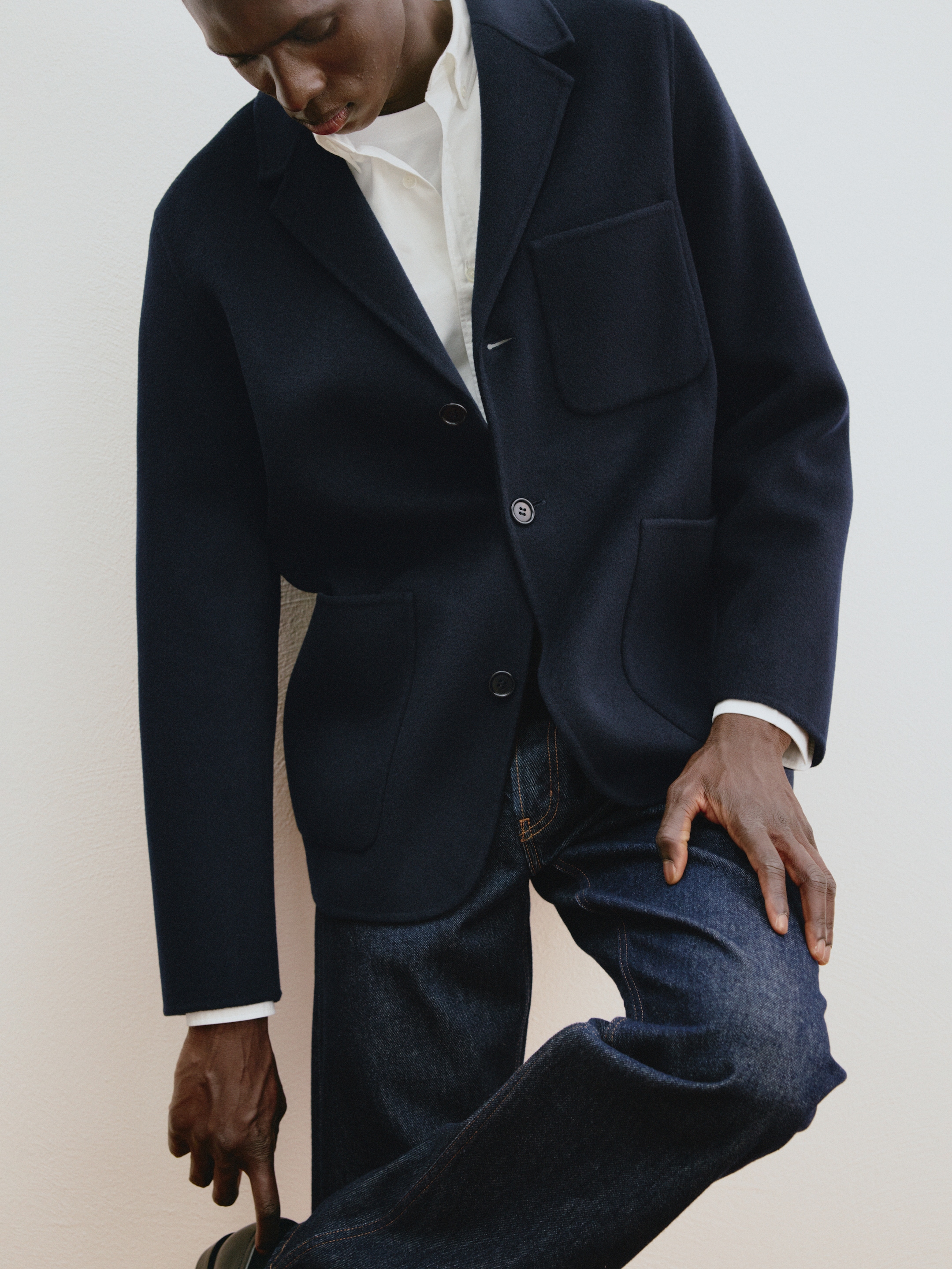 Person wearing a dark blazer, white shirt, and jeans, adjusting their shoe, standing against a light background.