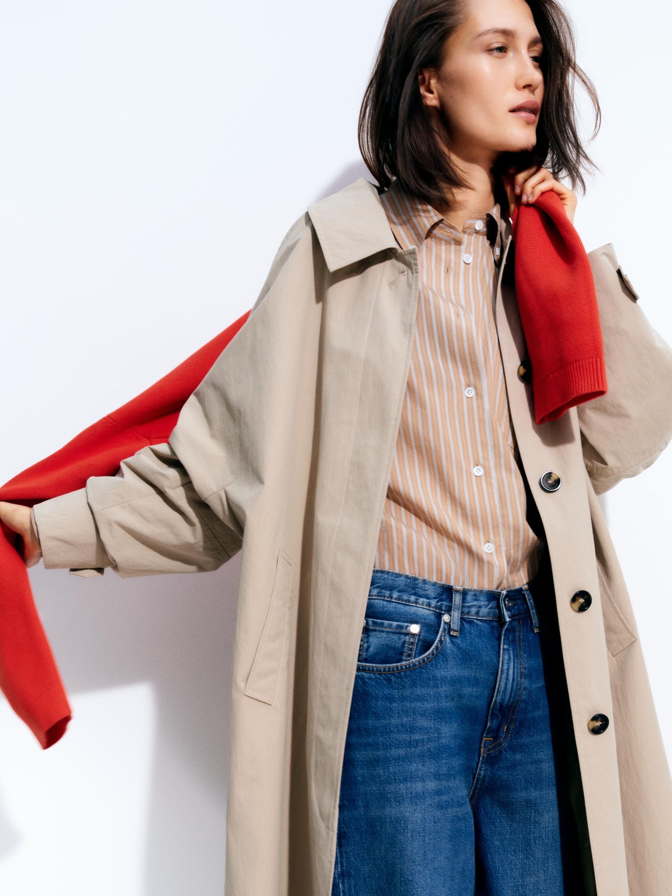 Woman in a beige trench coat over a striped shirt and jeans, holding a red sweater, stands against a white background.