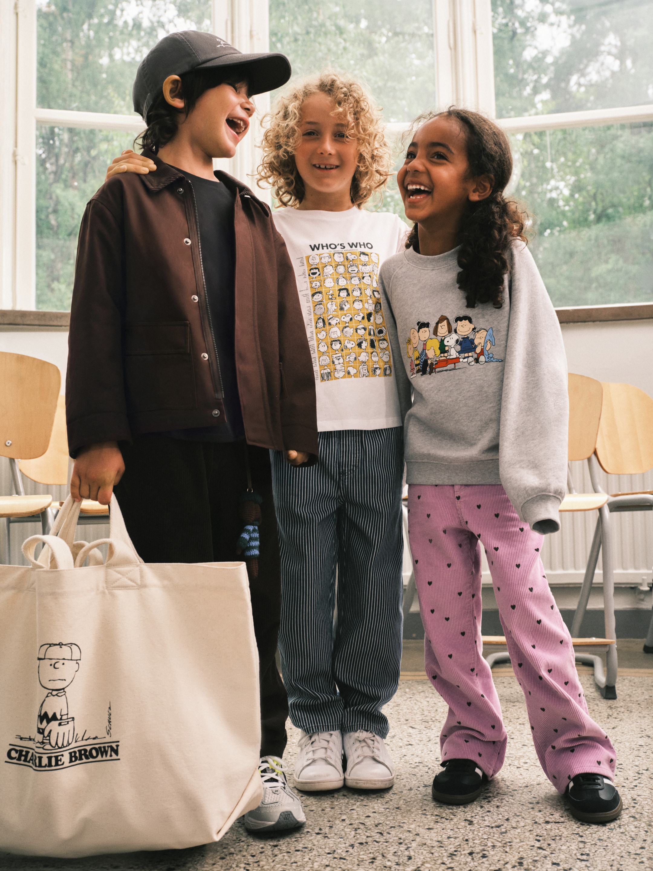 Three children smiling and standing together indoors, wearing Peanuts-themed clothing. One holds a bag with a Charlie Brown design.