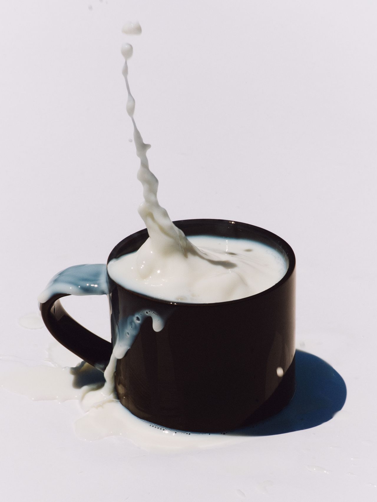 Milk splashing out of a black mug on a white surface, creating dynamic motion with droplets in mid-air.