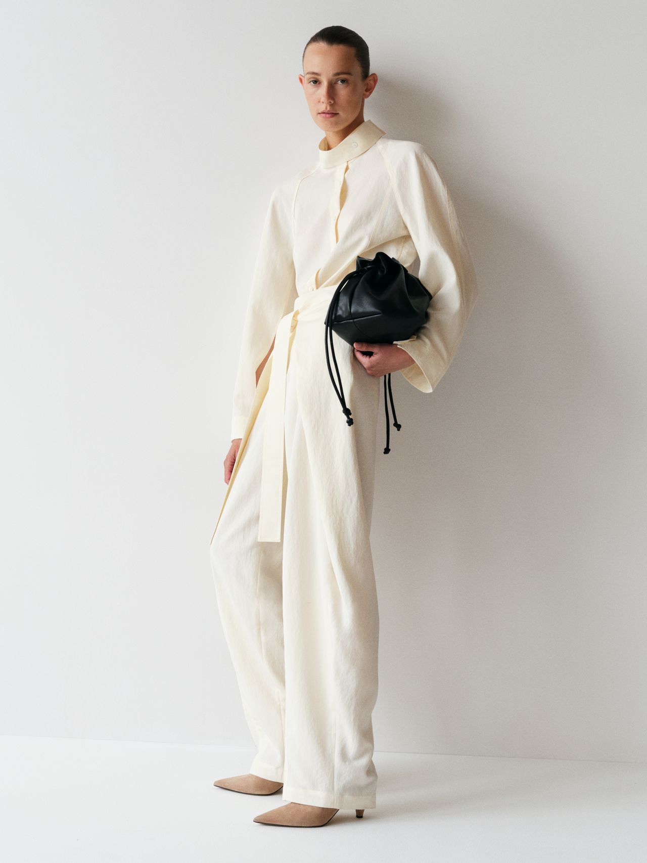 Person in a cream-colored outfit poses against a white wall, holding a black drawstring bag, wearing tan pointed shoes.