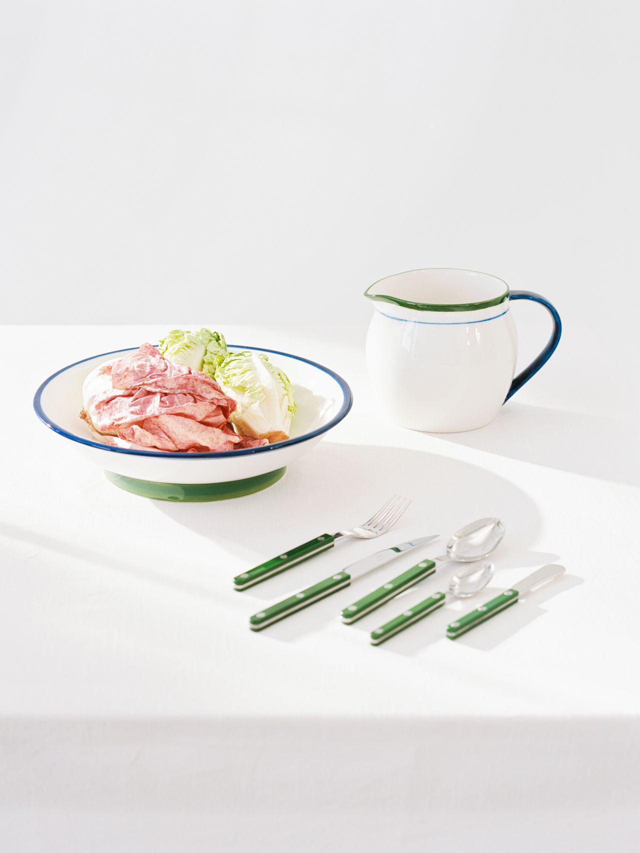 Plate of sliced meat and lettuce on a white tablecloth, with green-handled cutlery and a white pitcher with a green rim nearby.