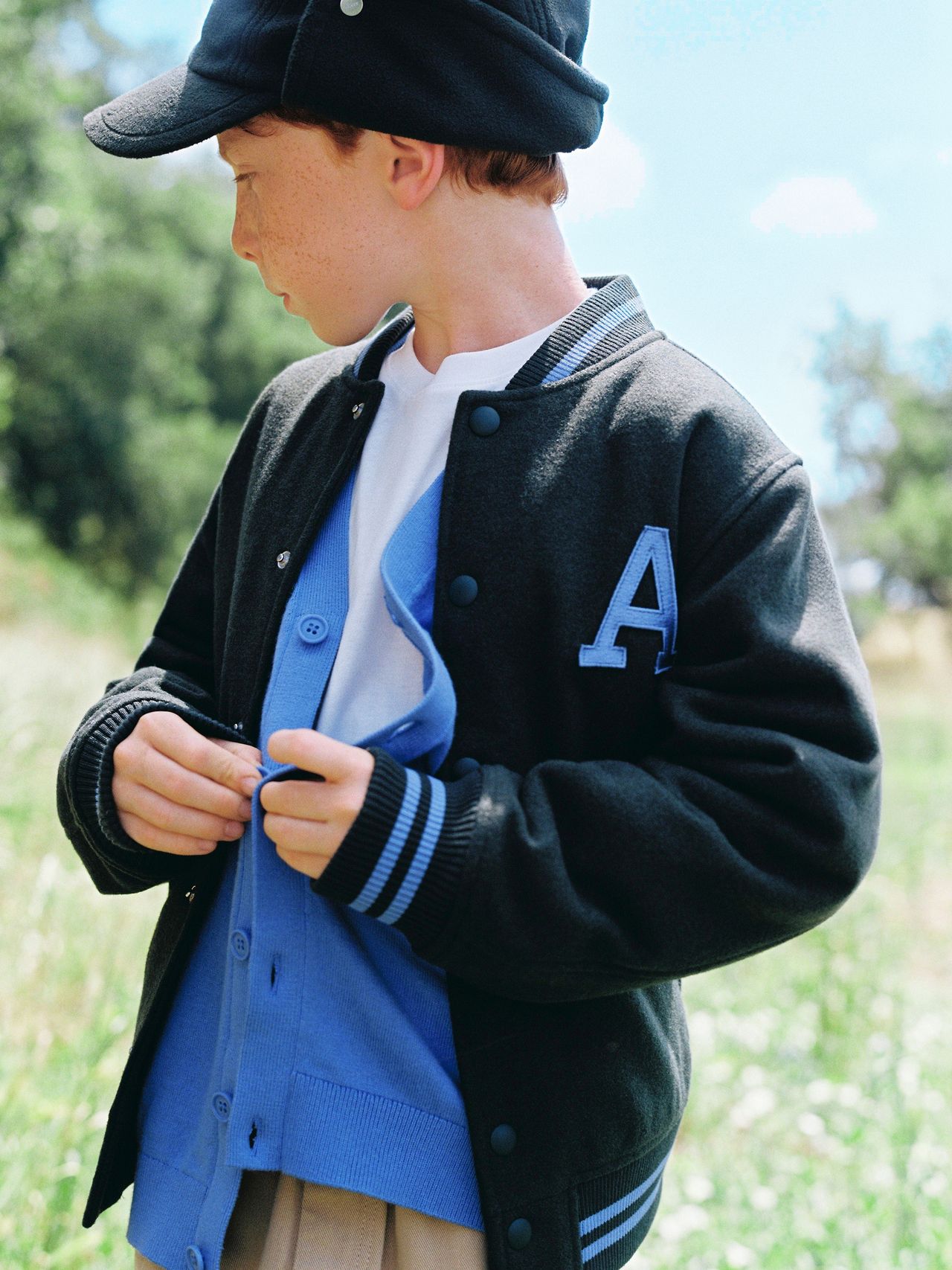 Young boy in a black cap and varsity jacket with a blue letter "A," standing outdoors, adjusting his blue shirt.
