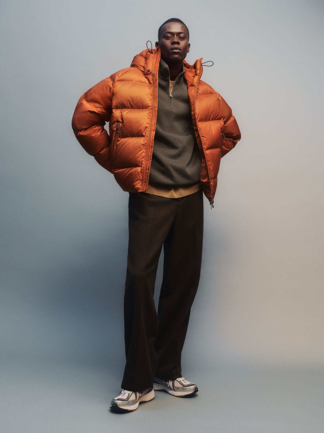 Person in an orange puffer jacket, dark pants, and sneakers stands confidently against a neutral background.